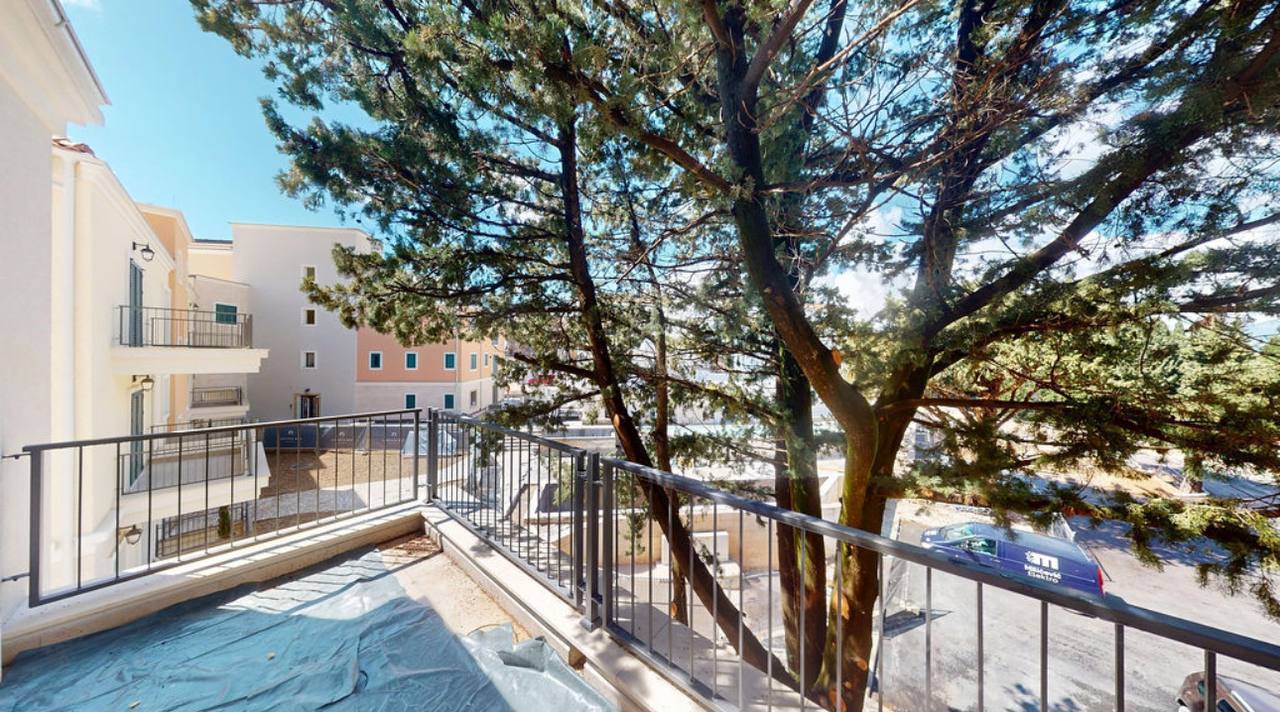 View from the apartment balcony in Lustica Bay, Montenegro: a modern building, wrought-iron railings, and a view of coniferous trees.