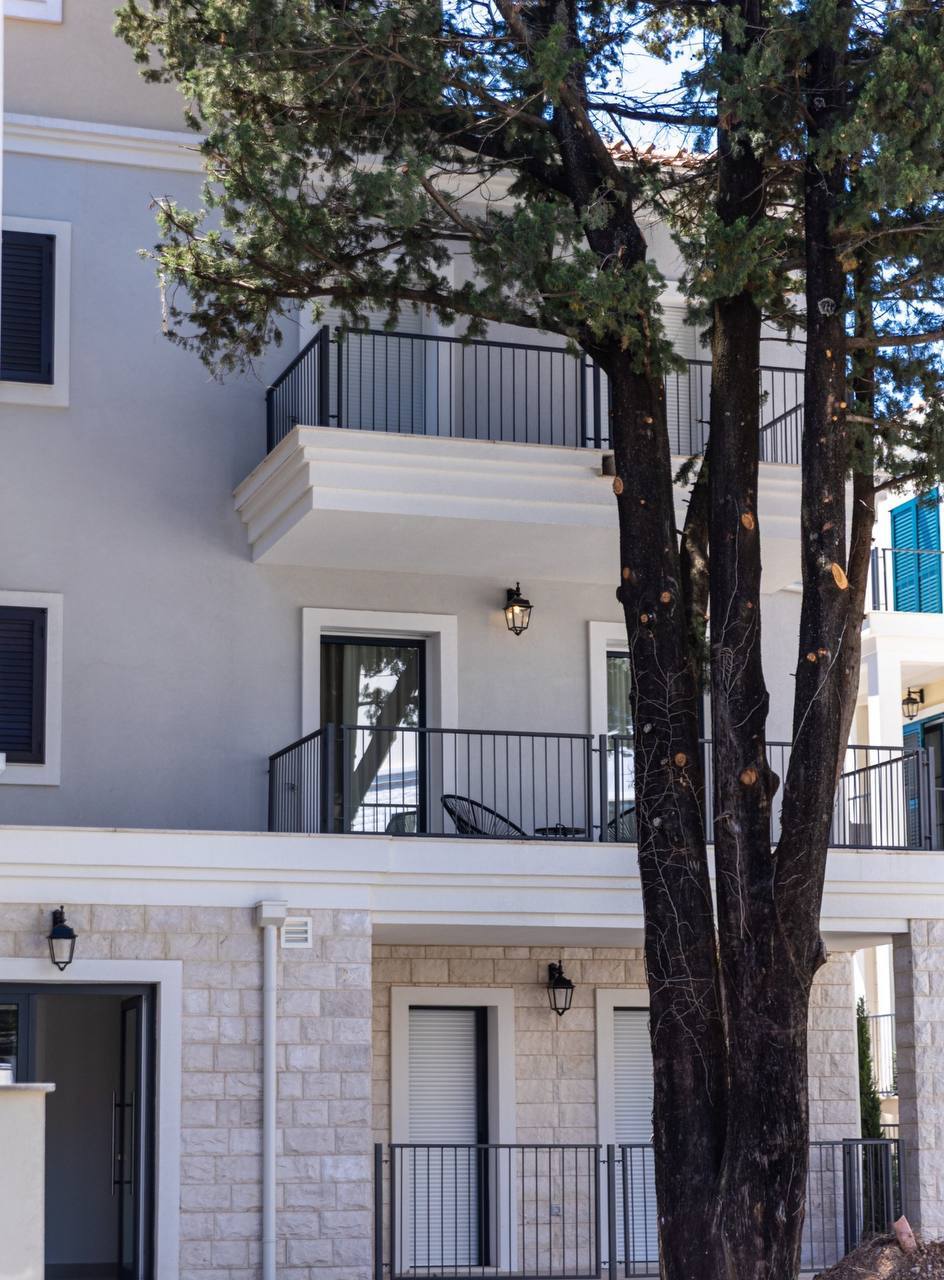 Facade of a modern apartment in Luštica Bay, Montenegro: light finish, stone base, balconies, and a view of cypress trees.