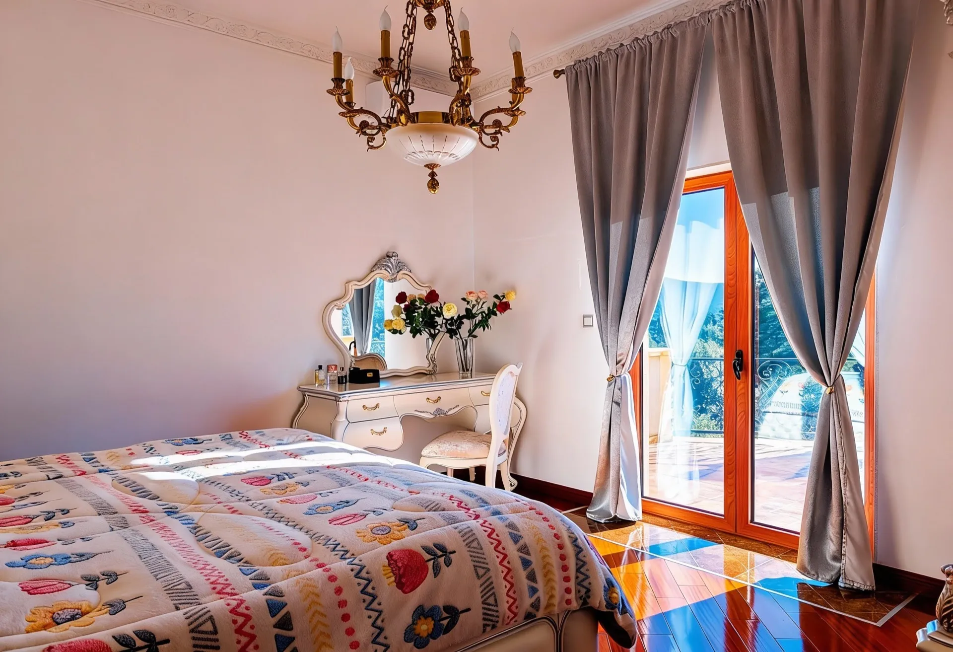 Bright bedroom with a colorful quilt, white vanity table, ornate mirror, and classic chandelier near a window with grey curtains.