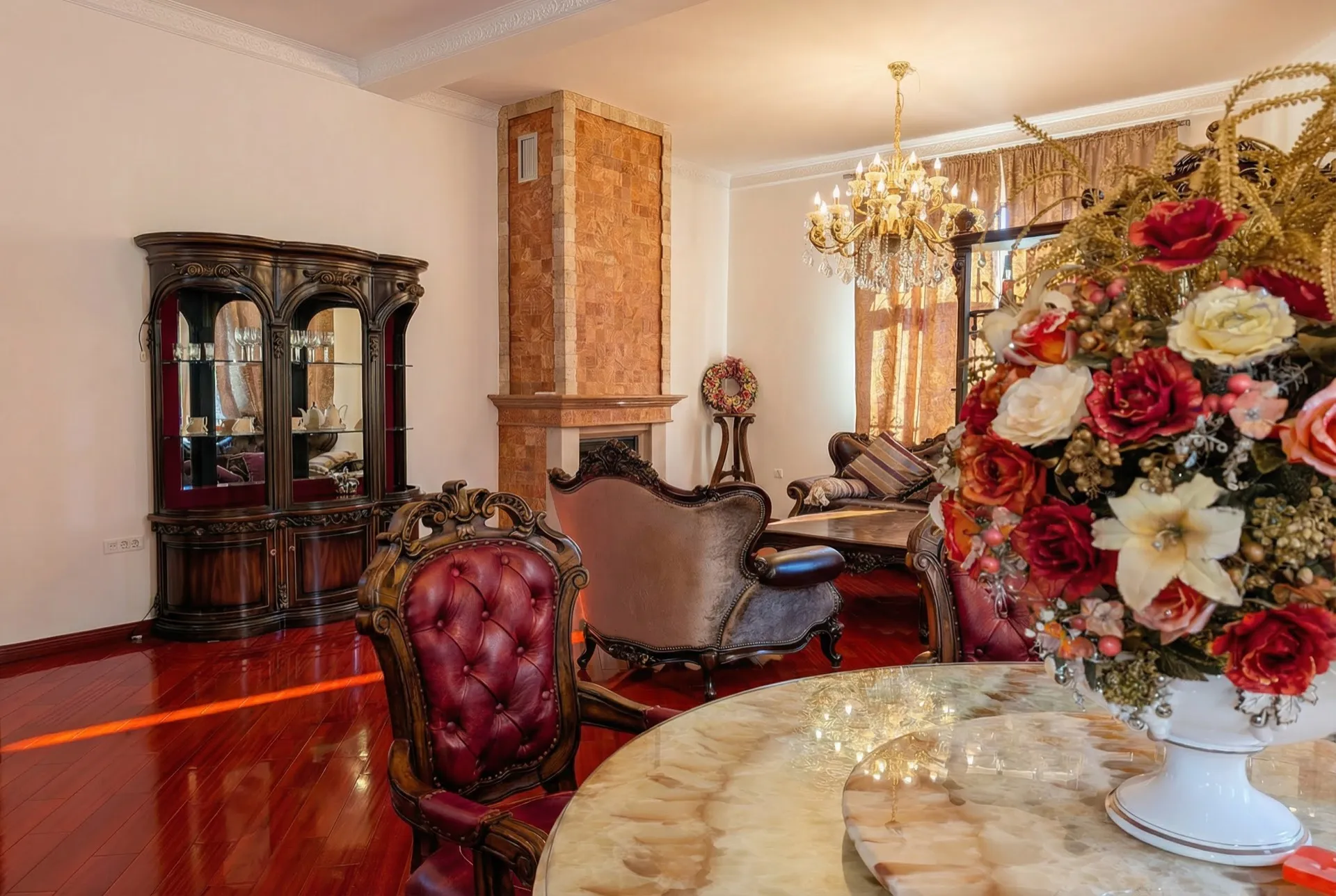 Luxurious classic living room featuring a stone fireplace, display cabinet, red leather armchairs, crystal chandelier, and polished hardwood floors.