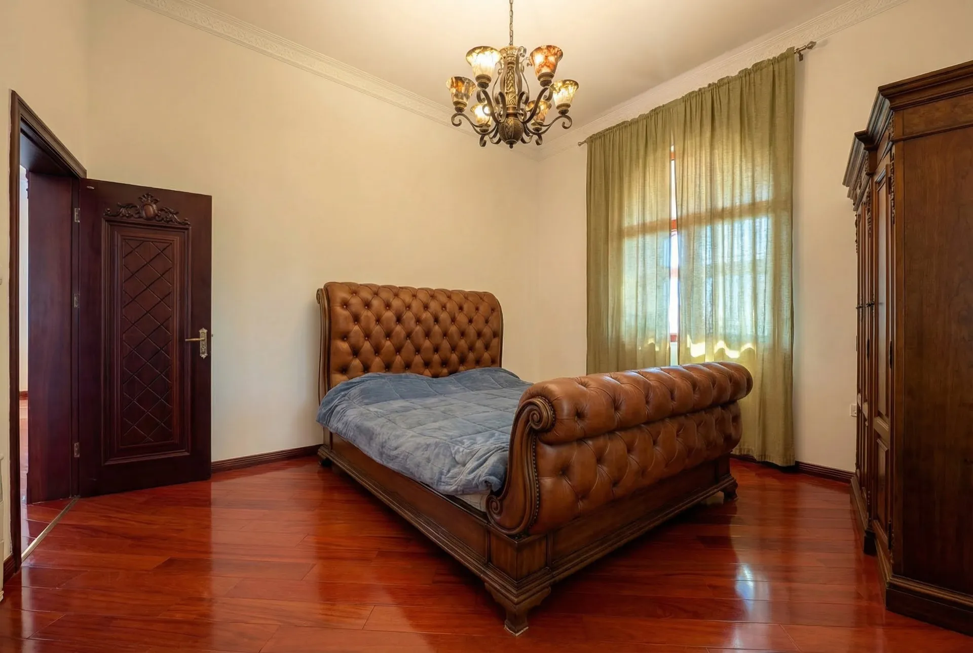 Bedroom featuring a large tufted leather bed, polished wood floors, an ornate chandelier, and green curtains.