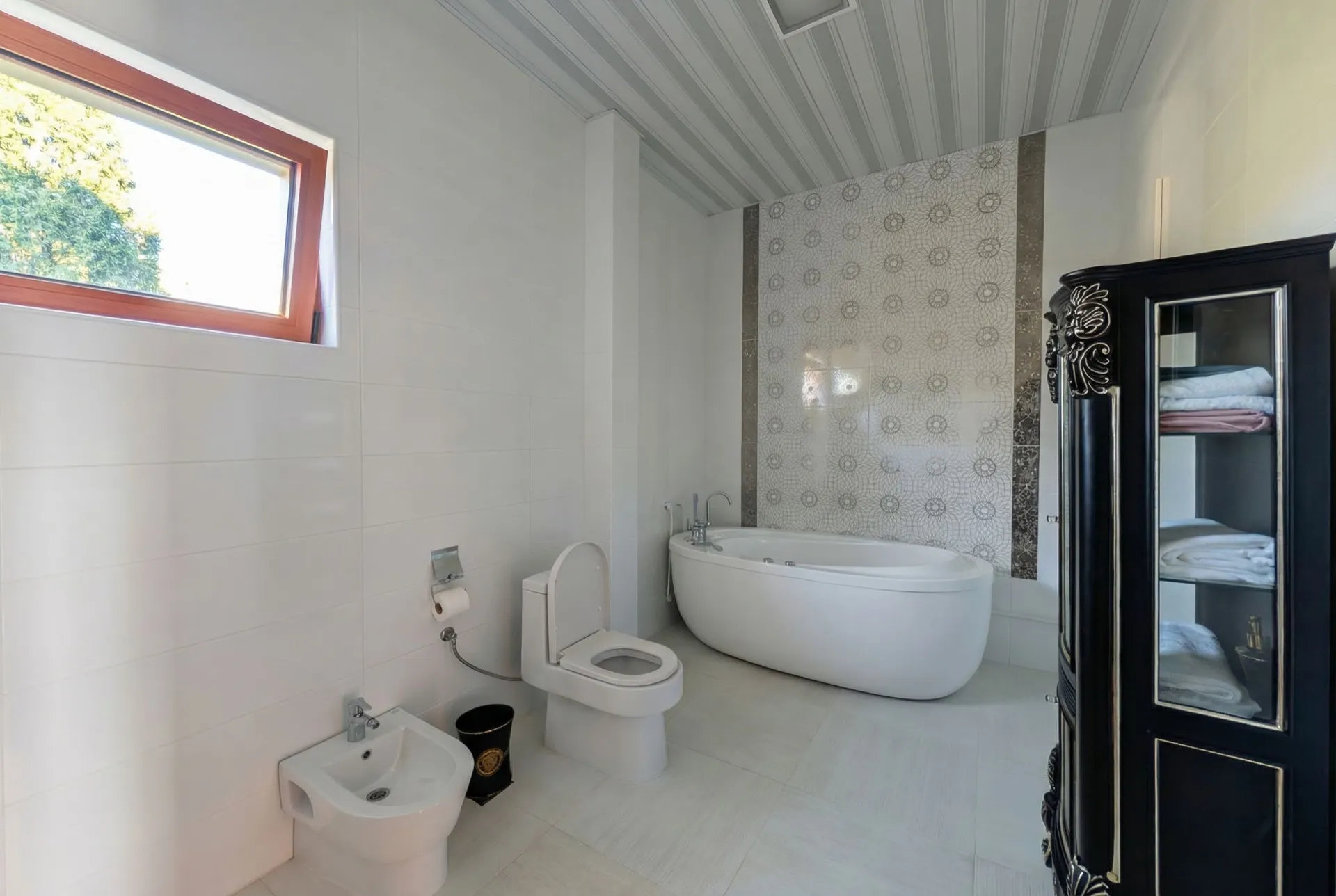 Bright bathroom with white freestanding bathtub, toilet, bidet, and dark cabinet for towels near a window.