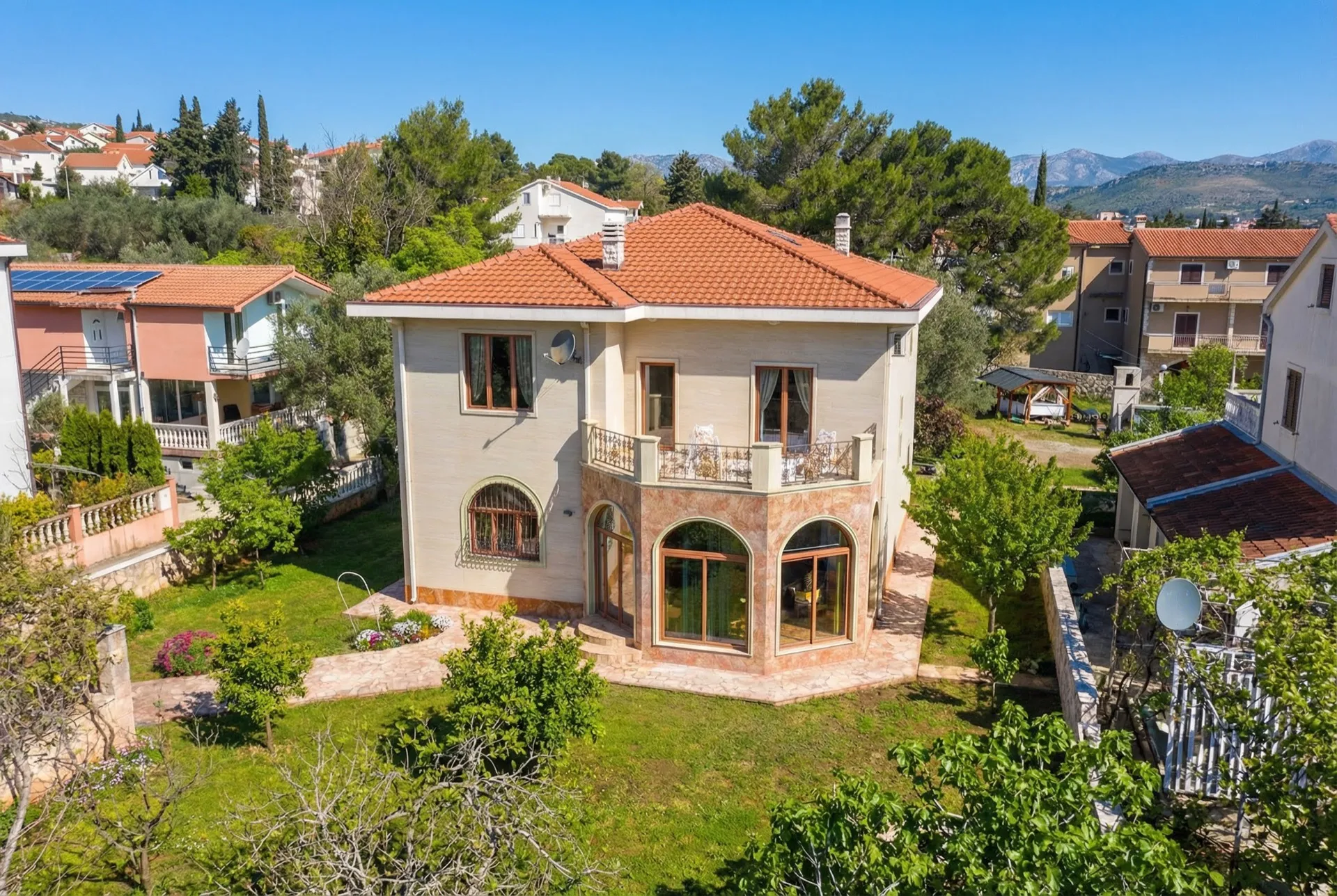 Aerial view of a two-story house with a red tiled roof and balcony, surrounded by a green garden and other houses against a mountain backdrop.