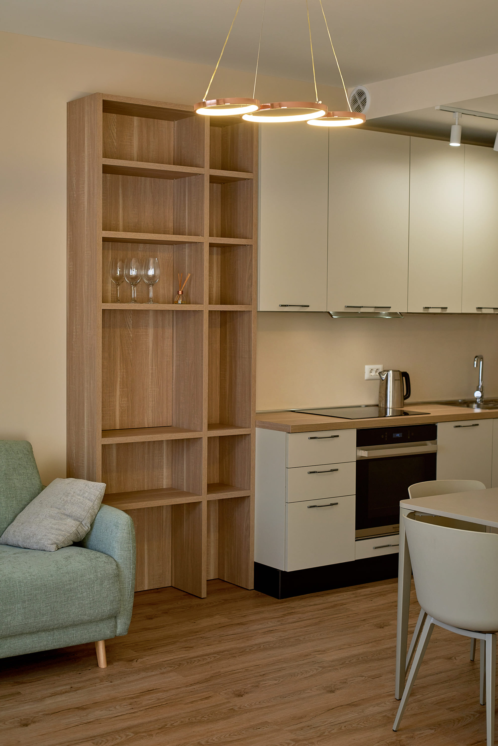 Modern kitchen-living room in an apartment, Bečići, Montenegro: light-colored kitchen set, wooden shelving unit, and a mint green armchair.