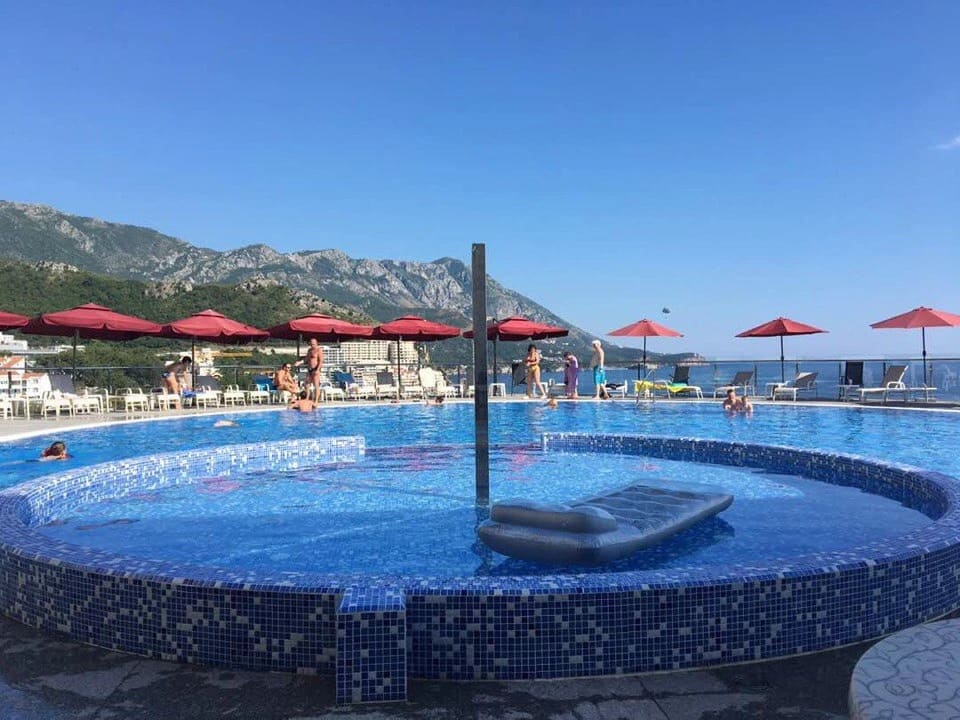 An outdoor swimming pool with a view of the sea and mountains in a residential complex in Bečići, Montenegro, with sun loungers and red umbrellas.