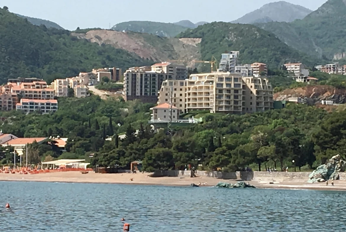 View of the residential complex and sandy beach in Bečići, Montenegro, against the backdrop of green mountains and the coastline.