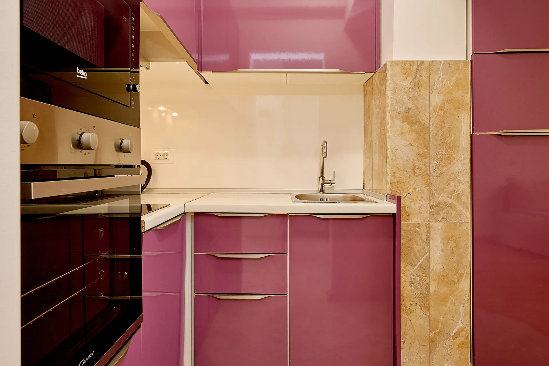Kitchen with glossy purple cabinets, built-in Beko and Candy appliances, white countertop, and a stainless steel sink.