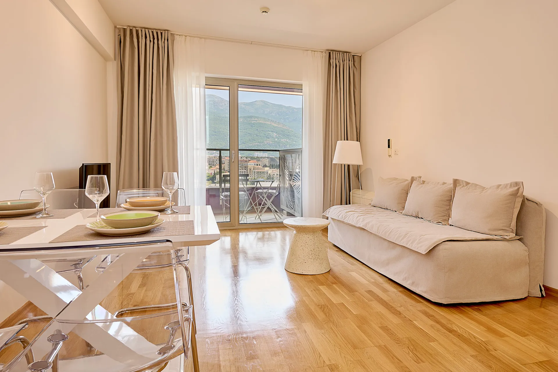 Bright living room with a beige sofa, white table, and transparent chairs. View of mountains through a large window.