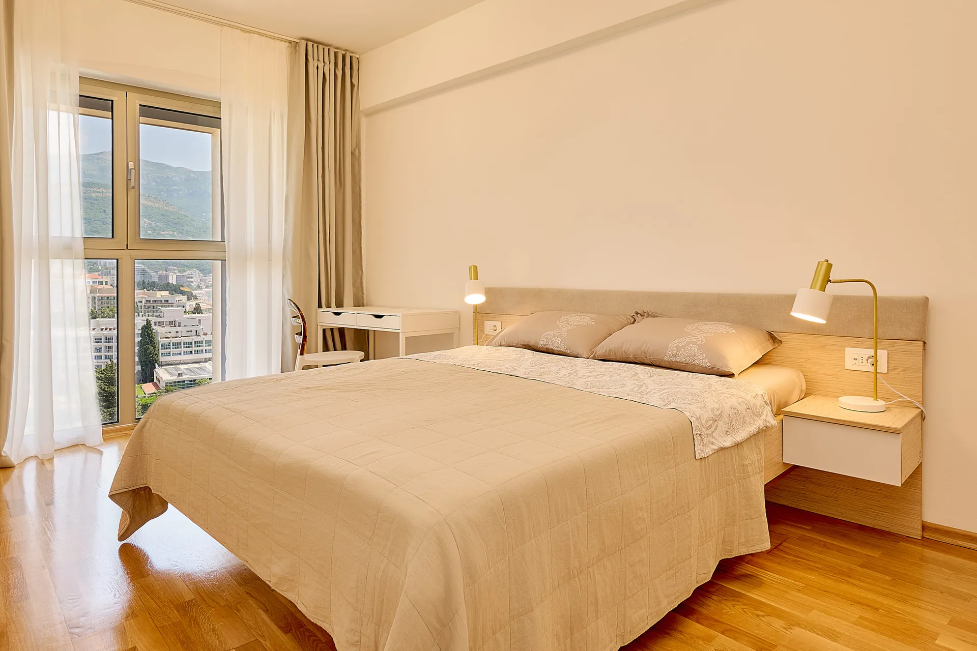 Bright bedroom with double bed, wooden floor, and window overlooking city buildings and green mountains.