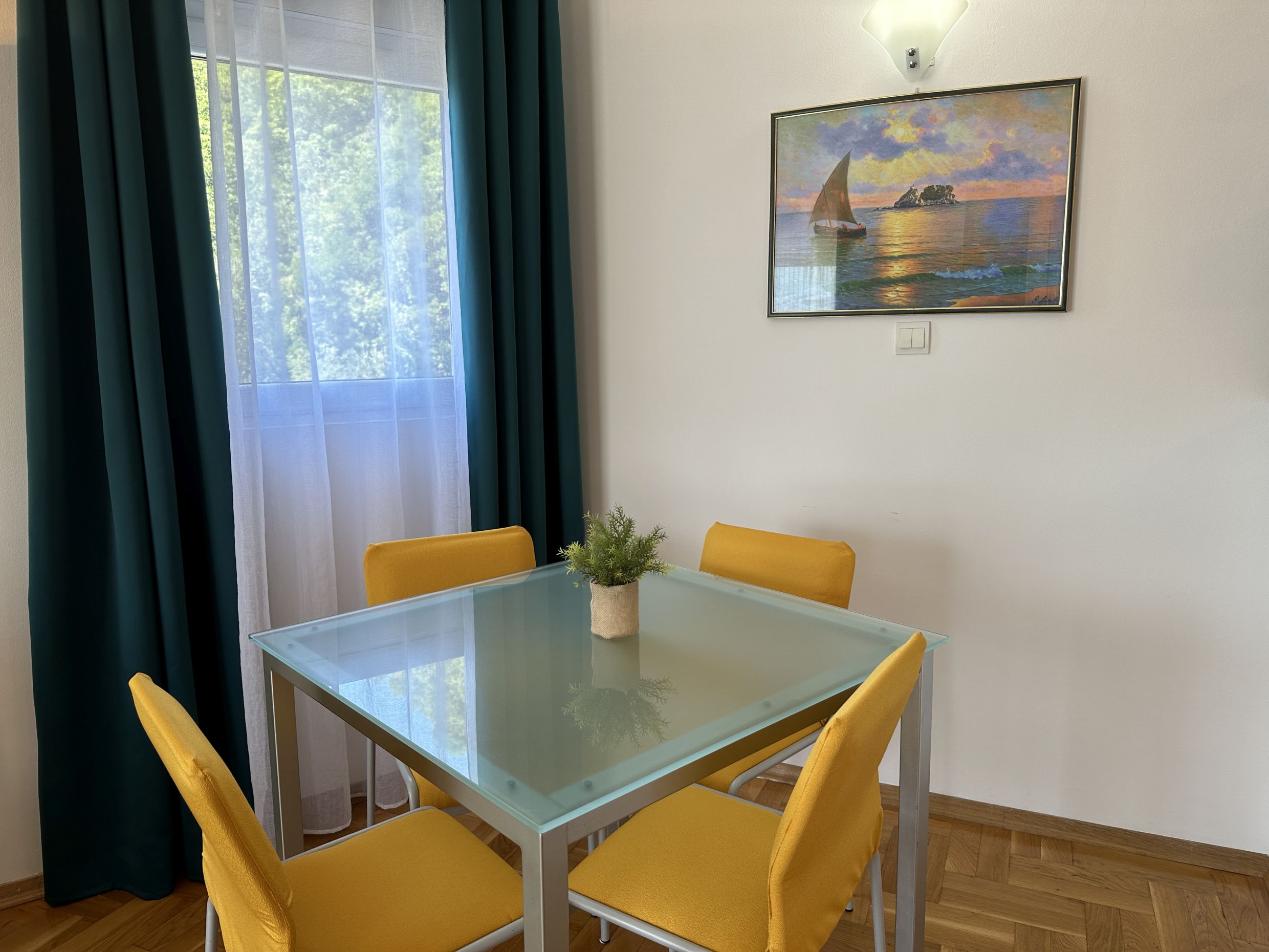 Dining area in the living room of an apartment in Bečići, Montenegro: a glass table, yellow chairs, and a view of the greenery.