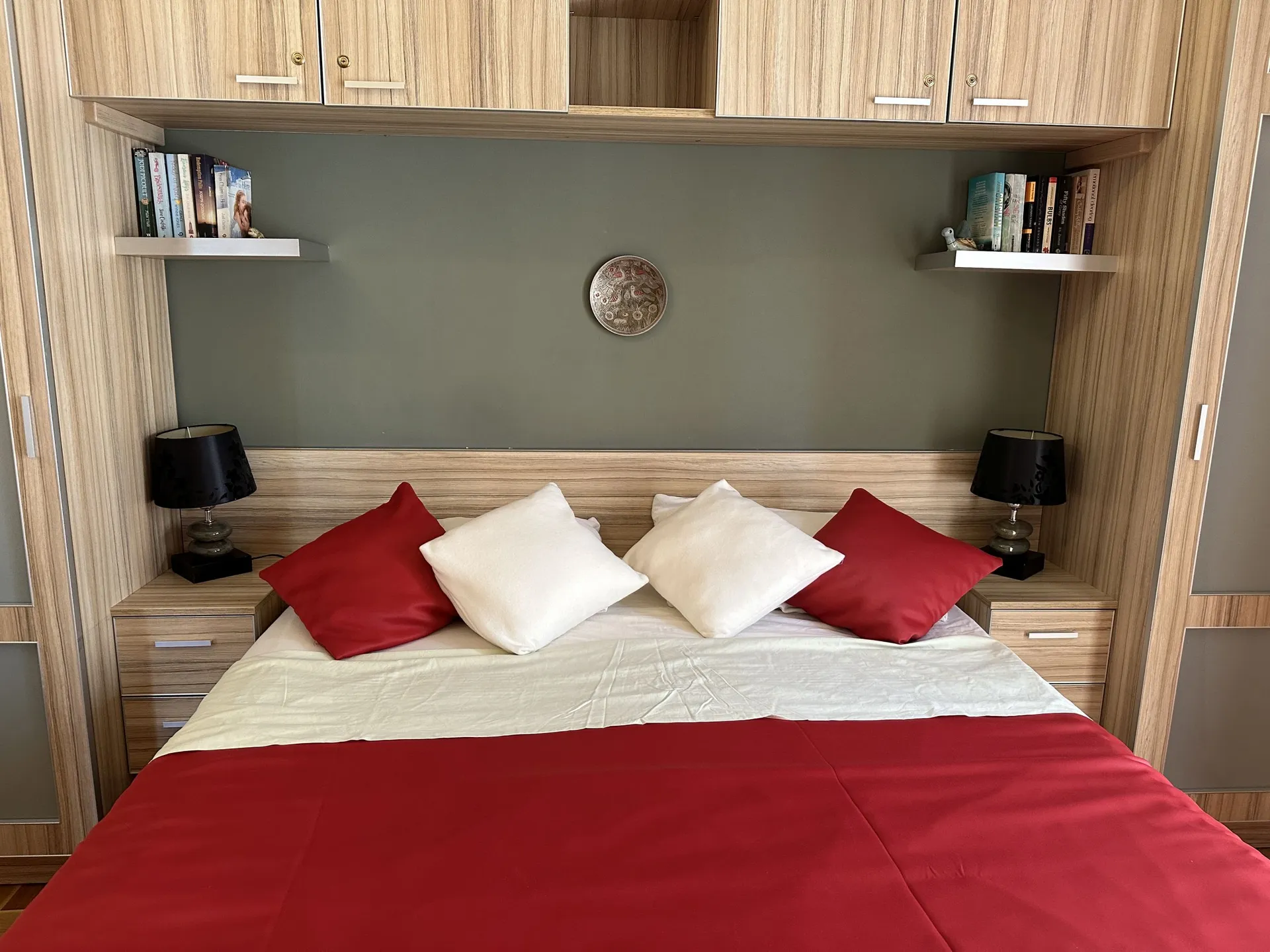 Bedroom featuring a bed with red and white bedding, wooden furniture, black lamps, and bookshelves mounted on a grey wall above the headboard.