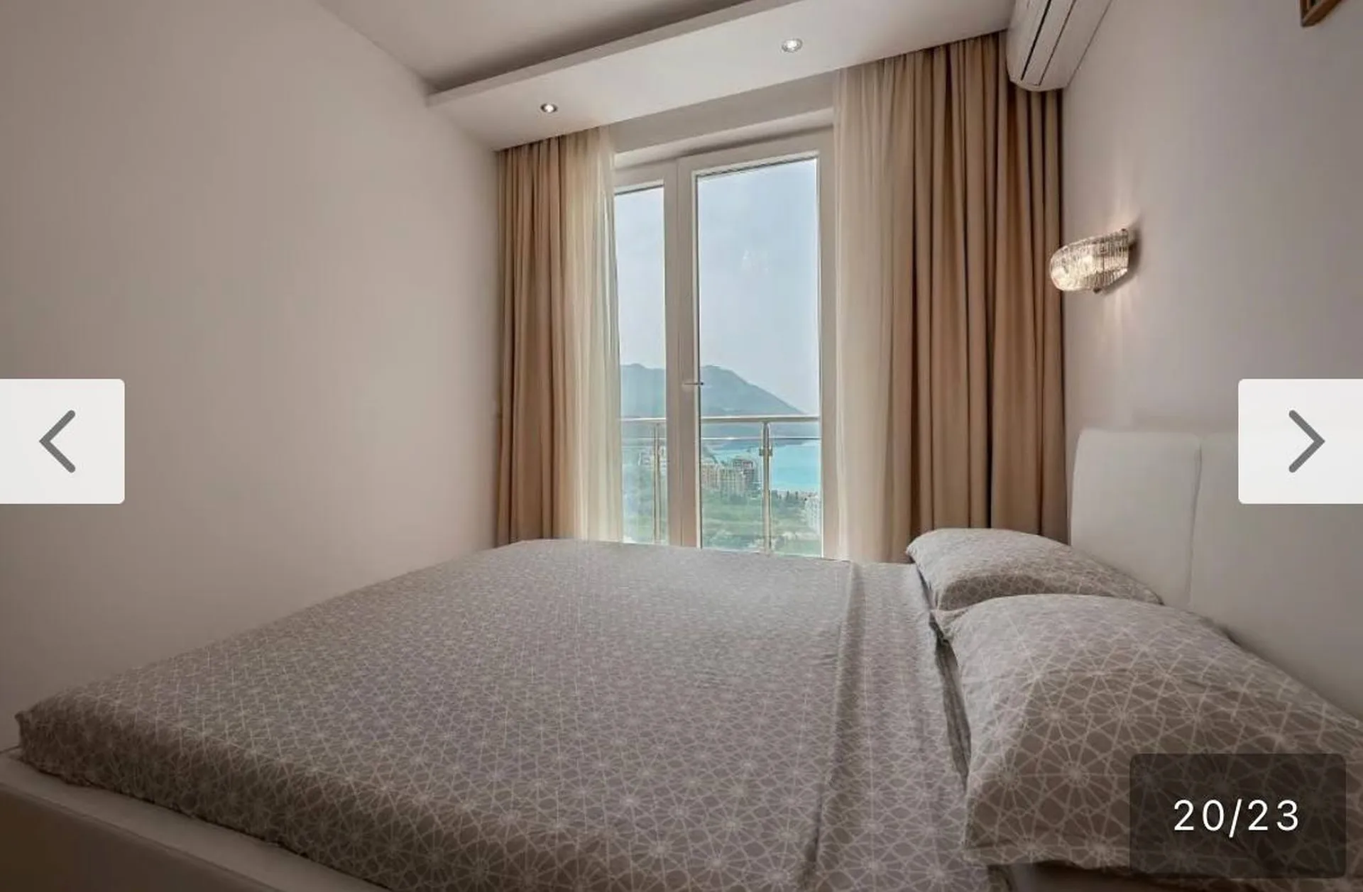 Bright bedroom with double bed and panoramic window. Sea and mountain view. Beige curtains, white headboard, air conditioner visible.