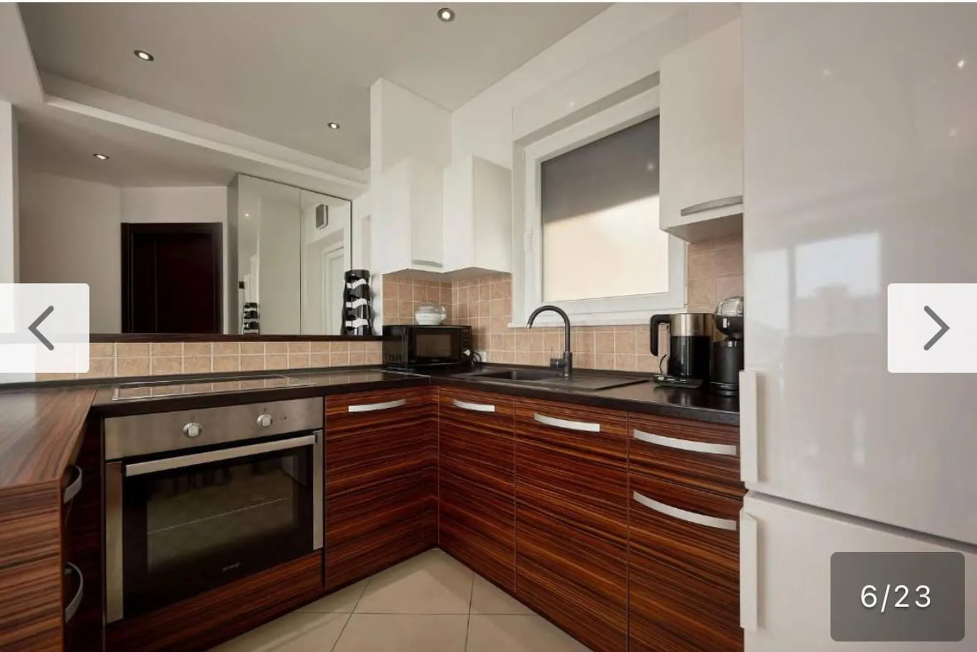 Modern kitchen featuring dark wood cabinetry, built-in oven, microwave, and white refrigerator near a window with beige tiles.