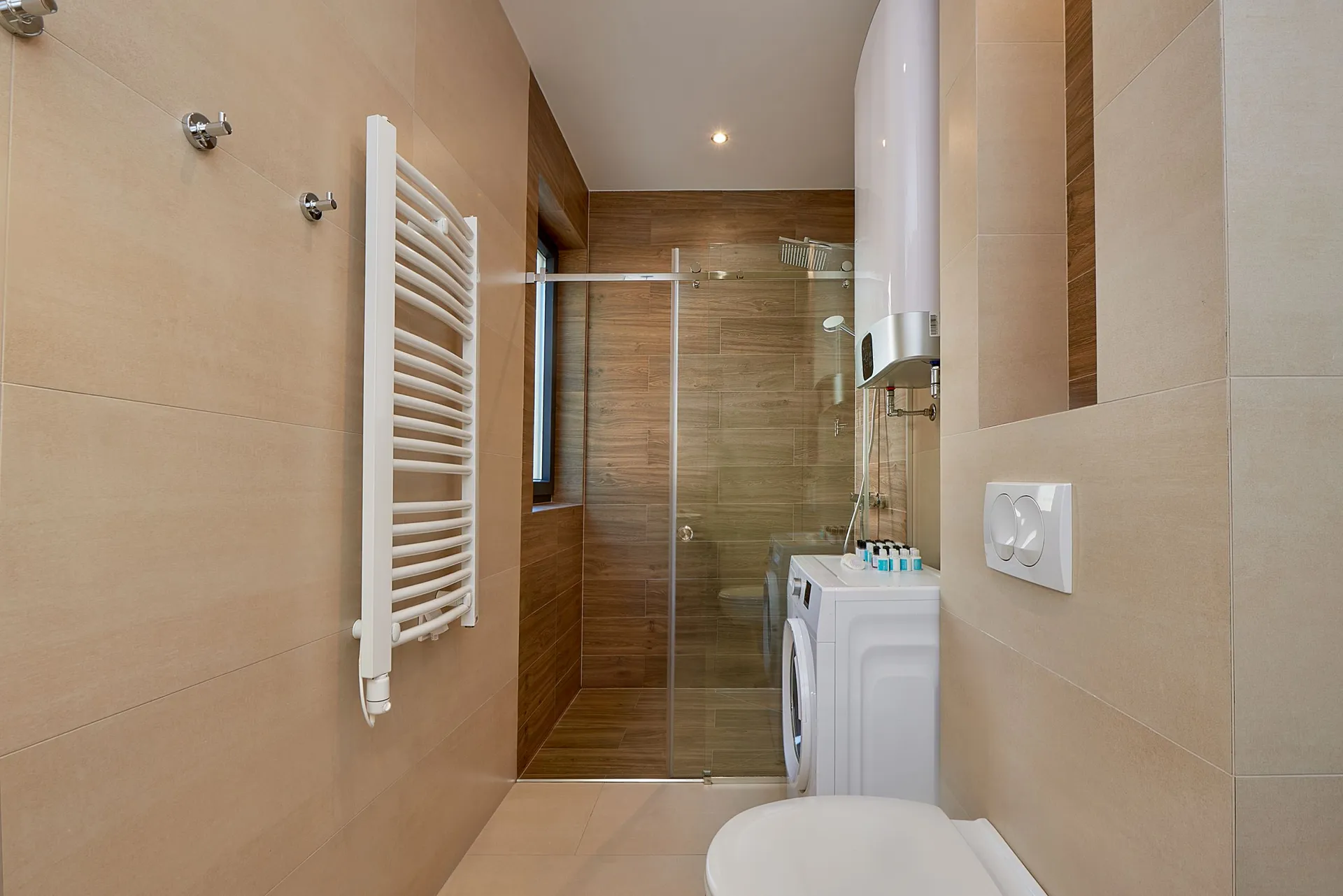Modern bathroom featuring a glass shower, washing machine, heated towel rail, and toilet against beige tiled walls.