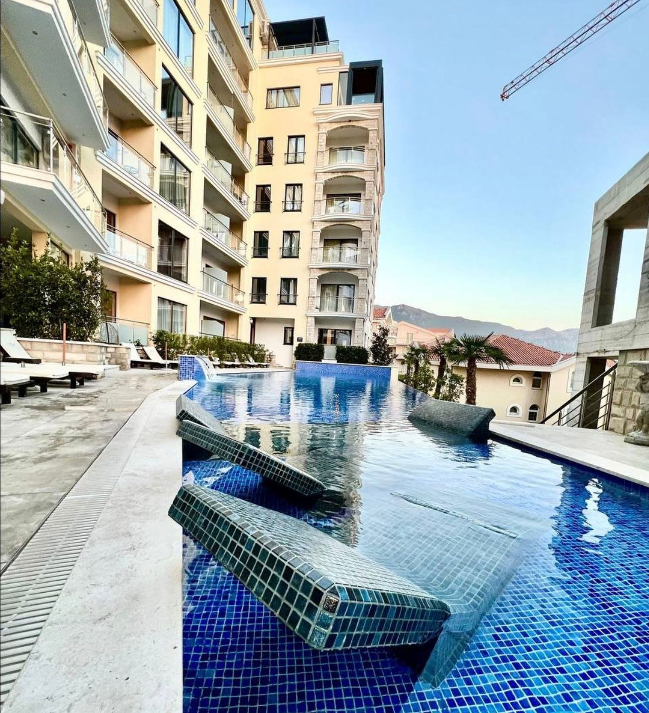 Outdoor pool with sun loungers in a modern residential complex in Bečići, Montenegro. View of a multi-story building and mountains.