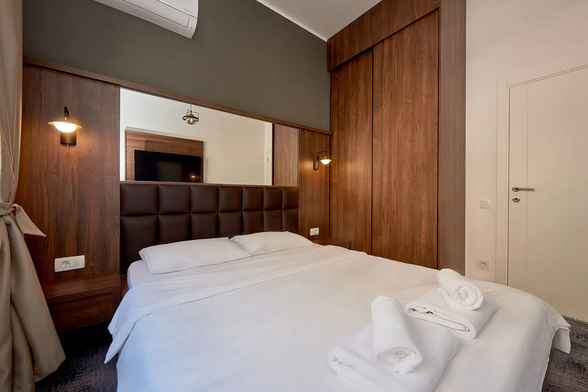 Modern bedroom with wooden paneling, a large mirror, and white bedding. Rolled white towels are placed on the bed.