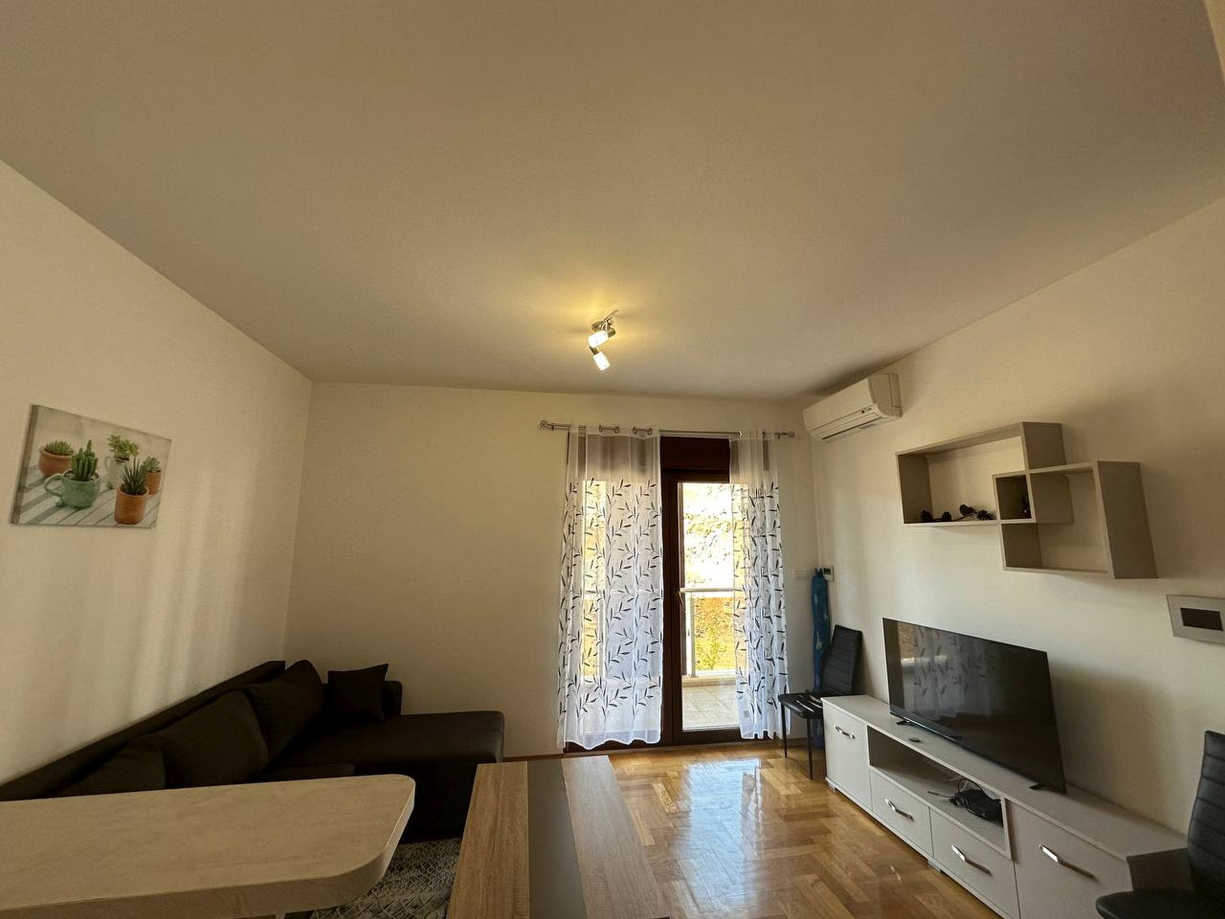 Bright living room with a sofa and TV area in an apartment in Bečići, Montenegro: parquet flooring, balcony access, modern furniture.