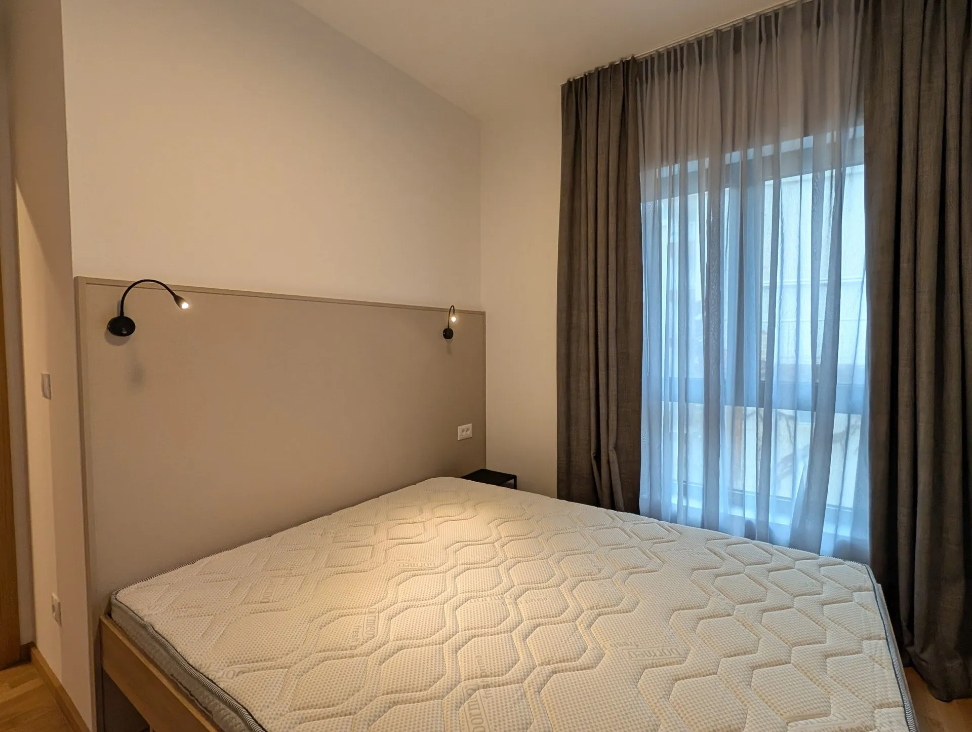 Modern bedroom with a white mattress, grey upholstered headboard, wall lamps, and a window covered by curtains.