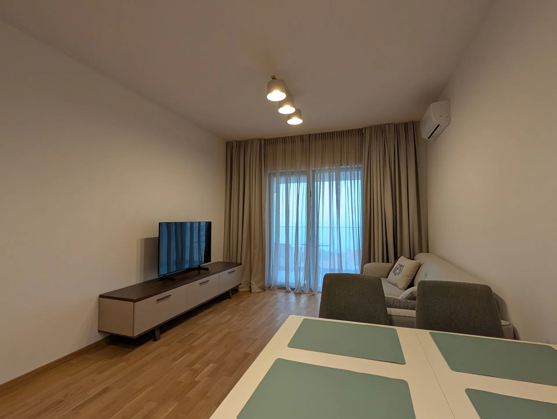 Bright living room with a sofa, TV on a console, and dining table near a window with beige curtains.