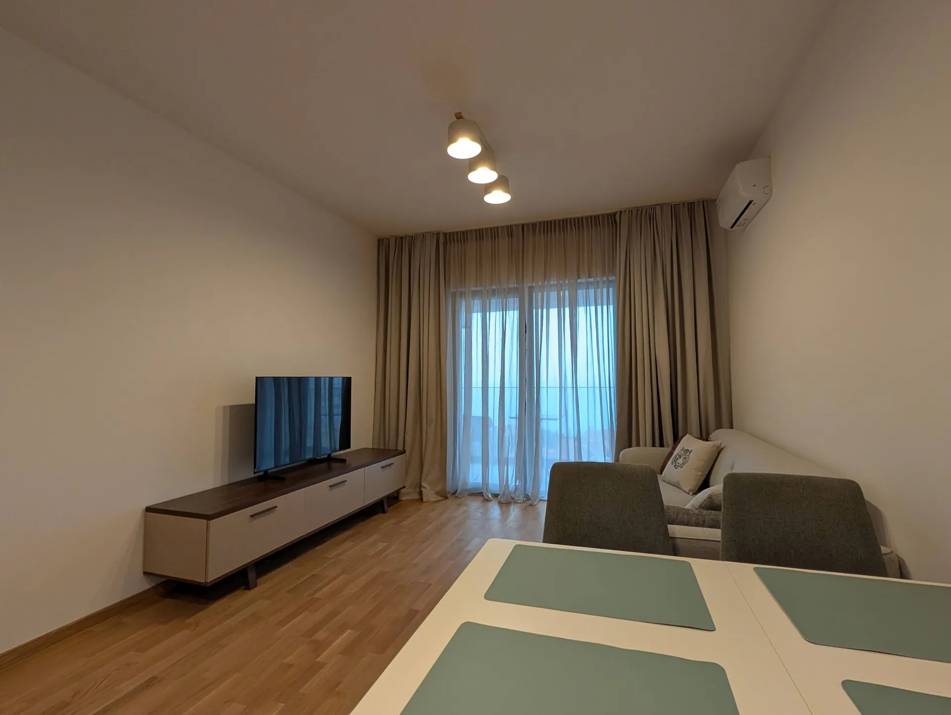 Bright living room with TV on a console, sofa, dining table, and large window covered by beige curtains.