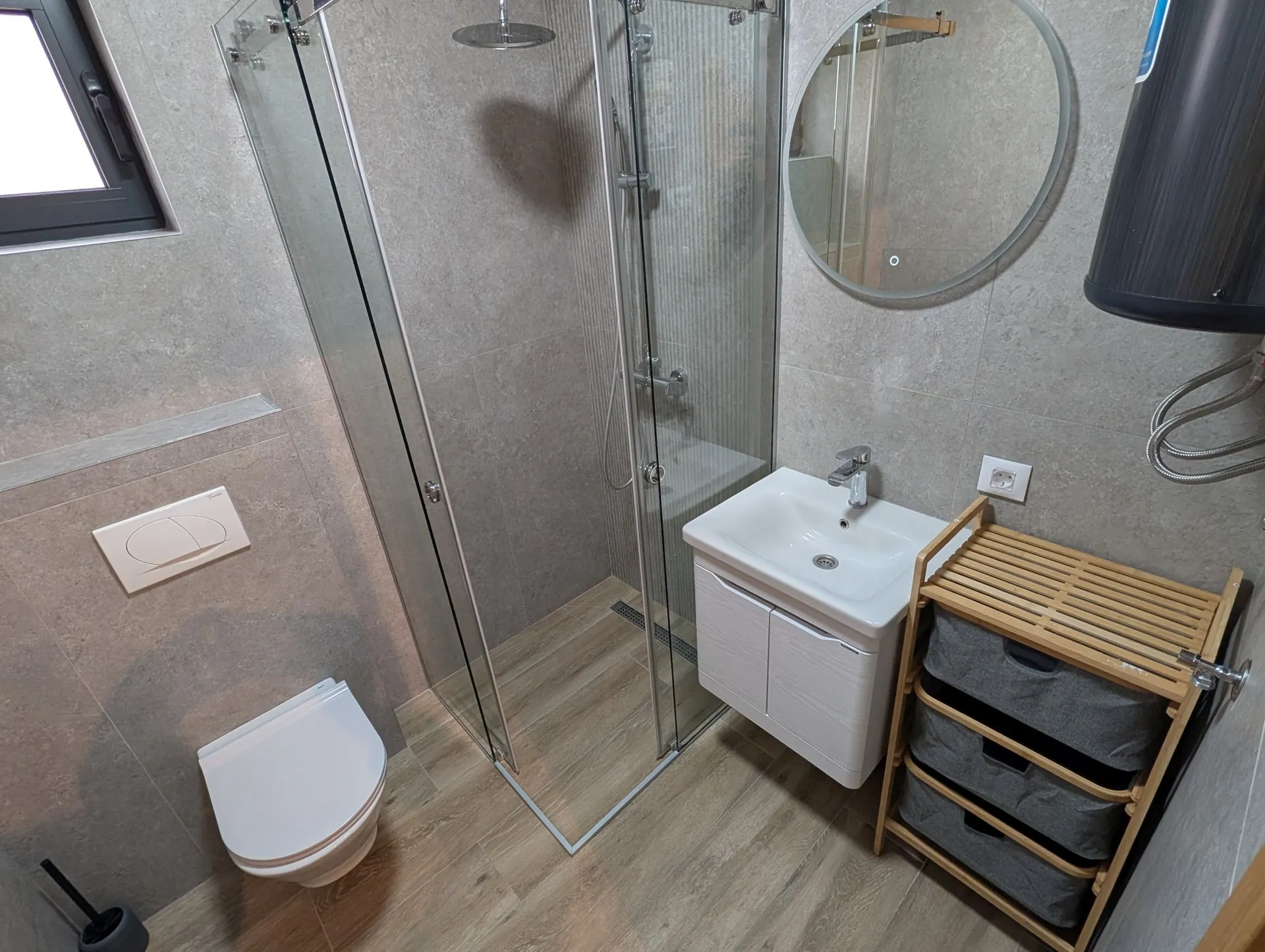 Modern bathroom featuring a glass shower, wall-mounted toilet, sink, and grey tiled walls.