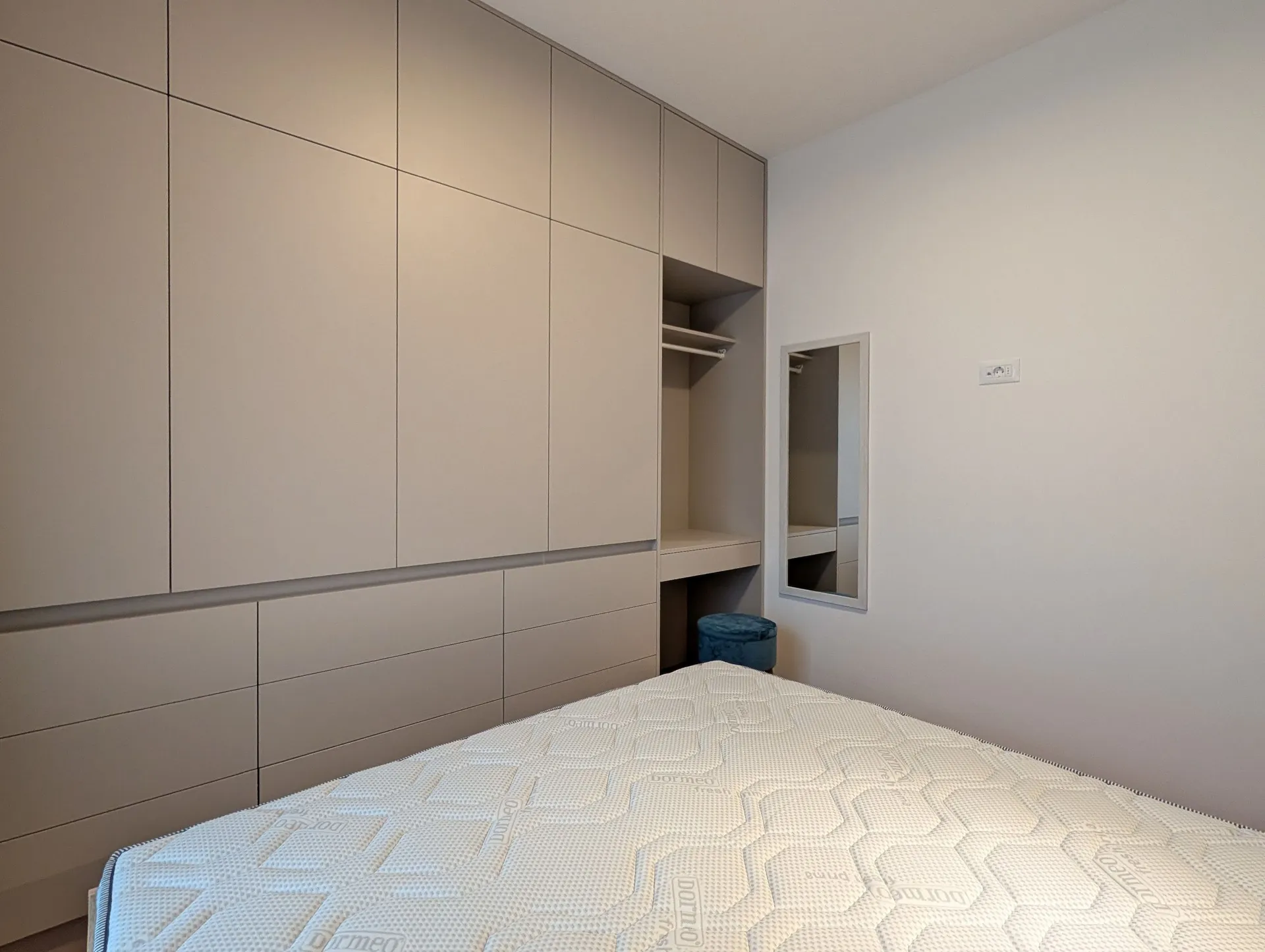 Modern bedroom featuring beige built-in wardrobe, mattress, and a small blue stool near the wall.