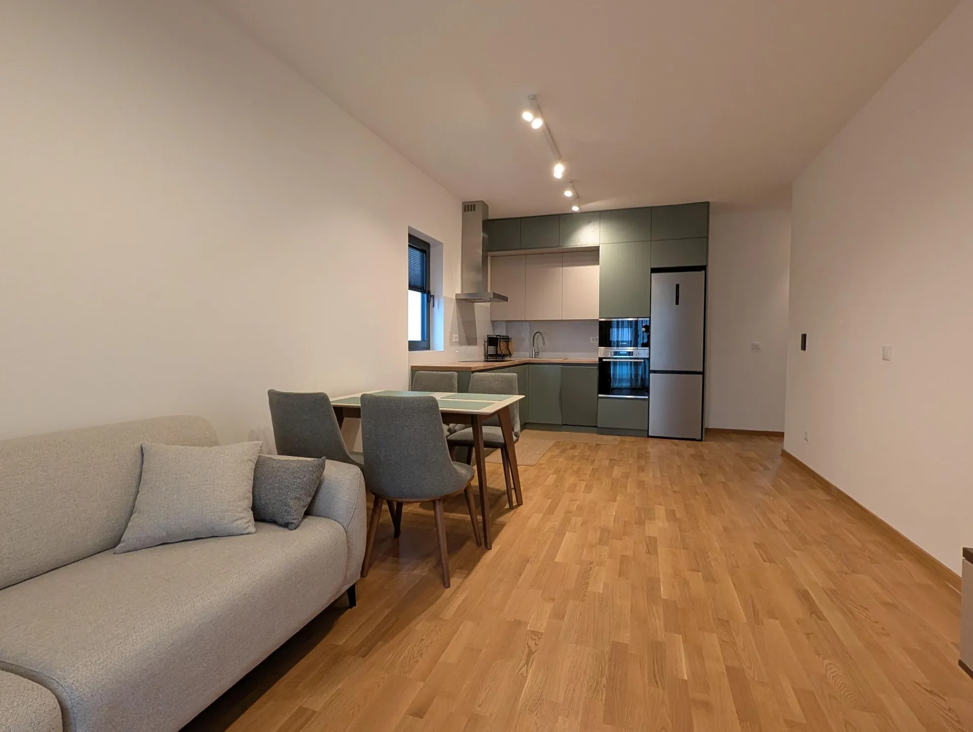 Modern studio apartment featuring a gray sofa, dining set, and green kitchen area on light wooden flooring.