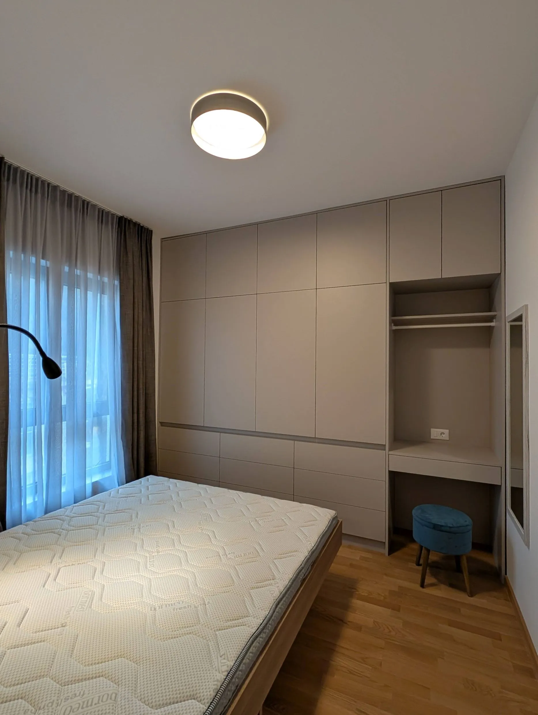 Modern bedroom with a mattress, built-in wardrobe, and blue stool near the window with curtains