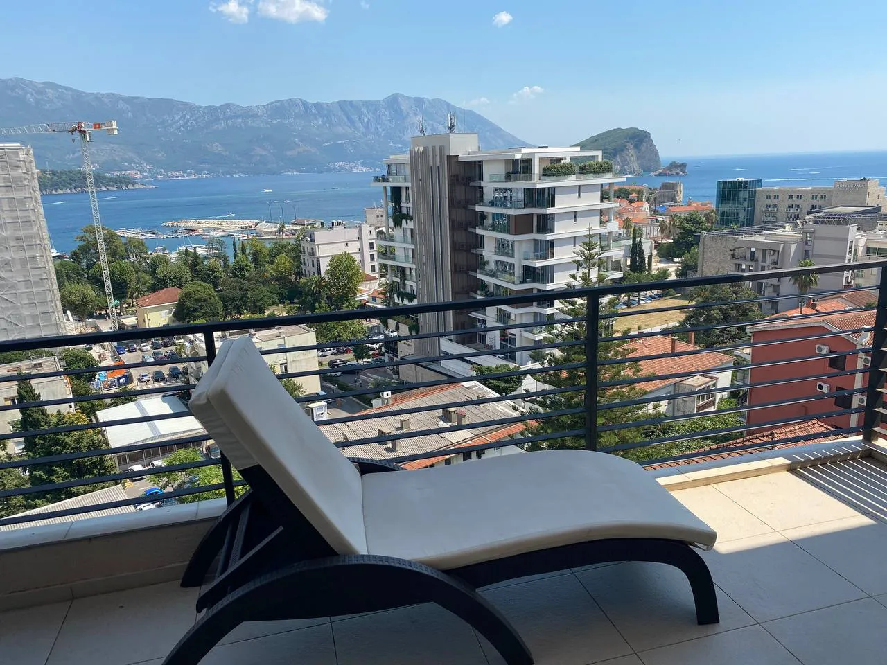 Balcony with a sun lounger overlooking the sea, mountains, and city buildings under a clear sky.