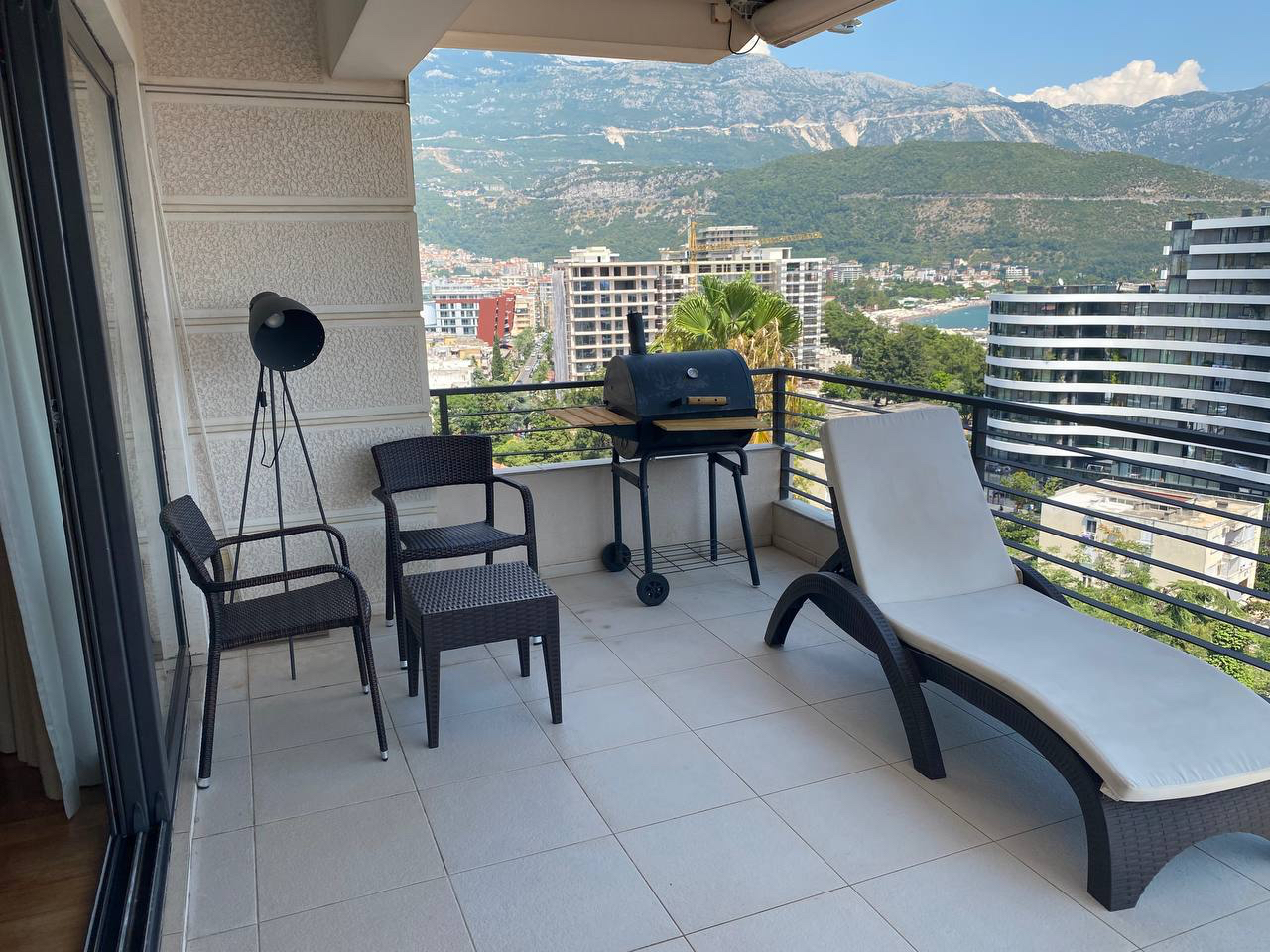 Apartment terrace in Budva, Montenegro: wicker chairs, a sun lounger, a grill, and panoramic views of the mountains and sea.