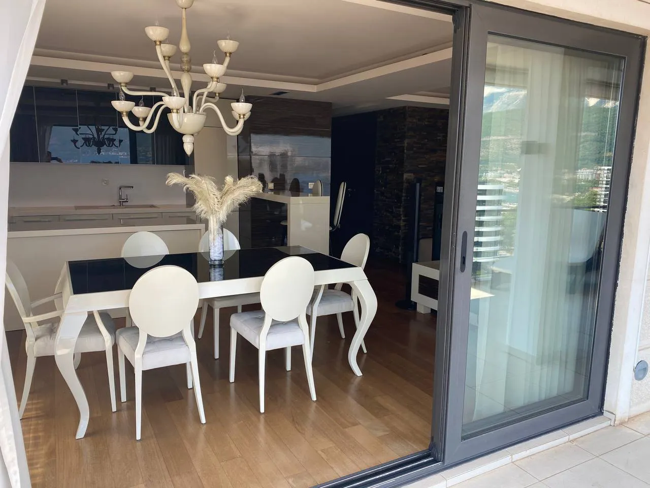 Bright living and dining area featuring a white table with six chairs and an ornate chandelier. A kitchen is visible in the background next to large glass sliding doors.
