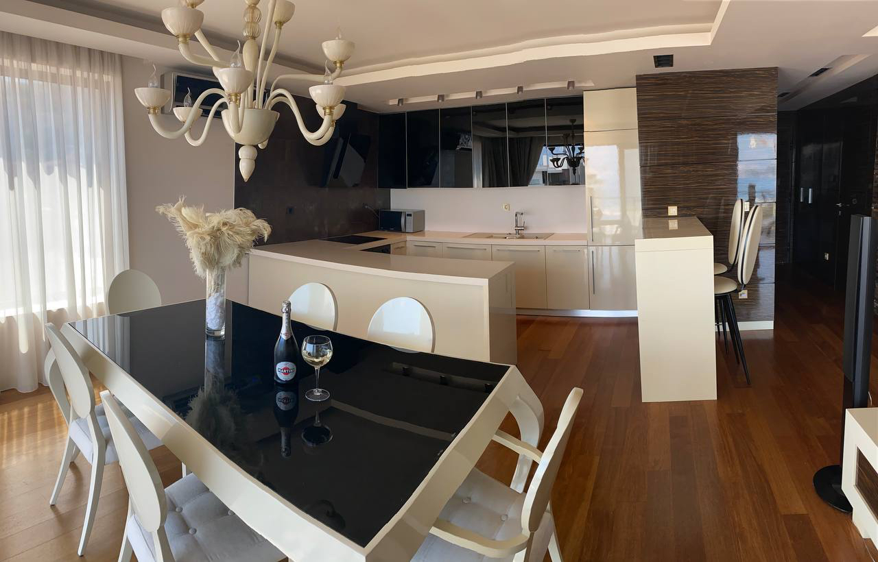 Modern kitchen-living room with dining area in an apartment in Budva, Montenegro: glossy fronts, a bar counter, and parquet flooring.