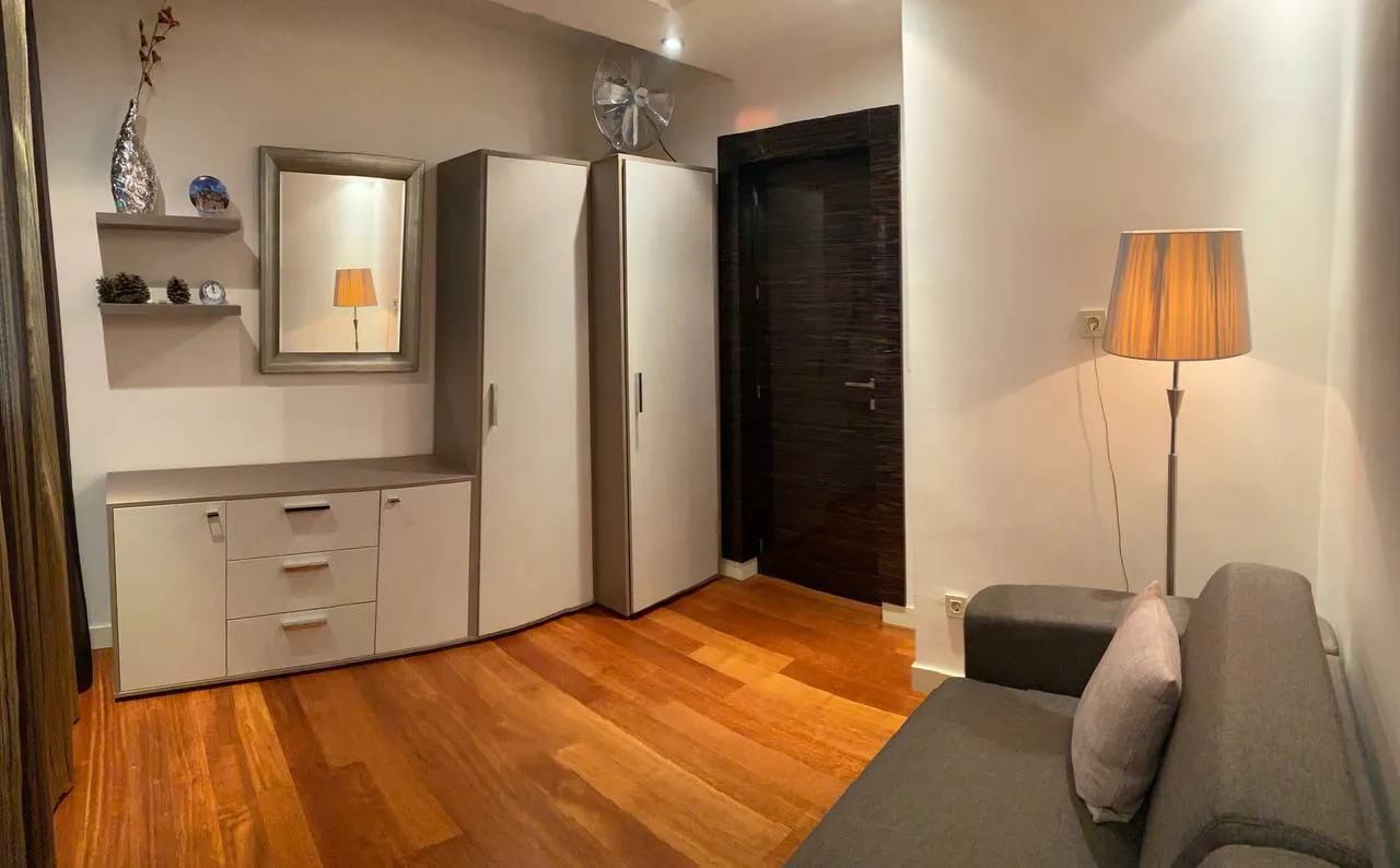 Living room with a grey sofa, light wardrobes, and wooden floor. Features a floor lamp, mirror, and dark entrance door.