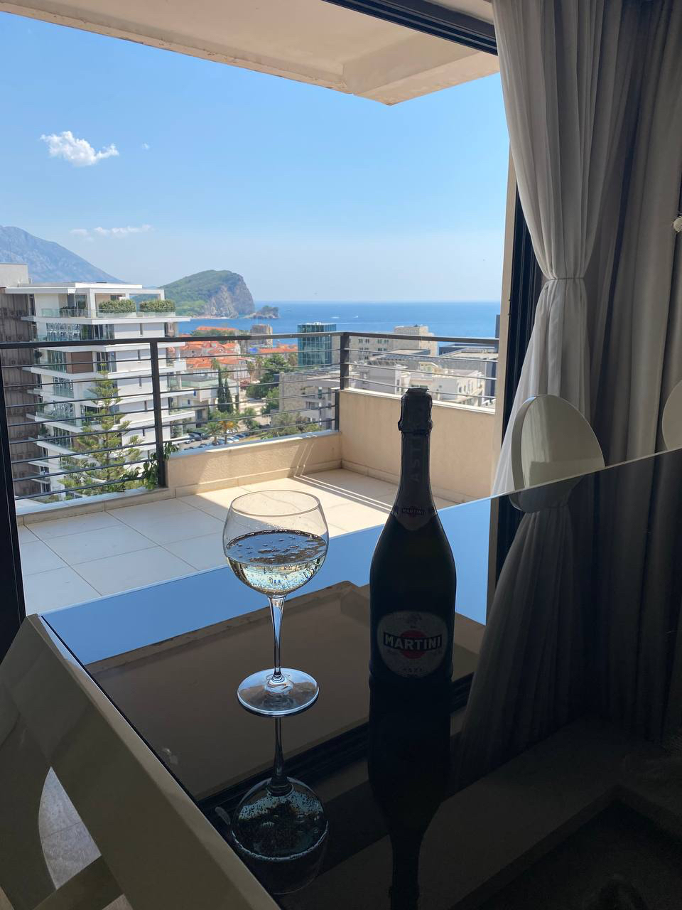 View from the apartment balcony in Budva, Montenegro: panoramic sea, mountain, and city views, with a glass table, a wine glass, and a bottle of wine.