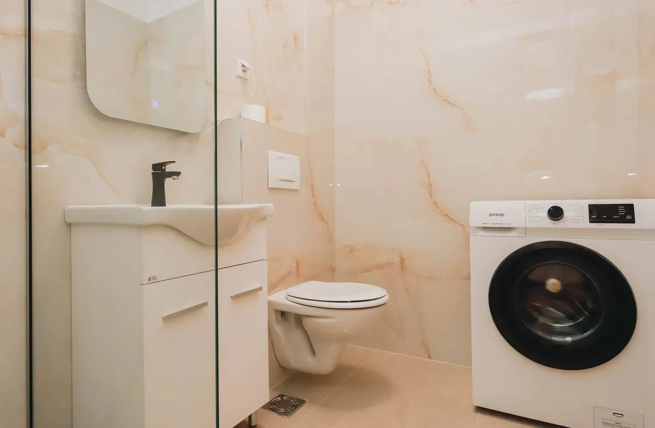 Modern bathroom featuring a white vanity sink, wall-mounted toilet, and washing machine set against beige marble-effect tiled walls.