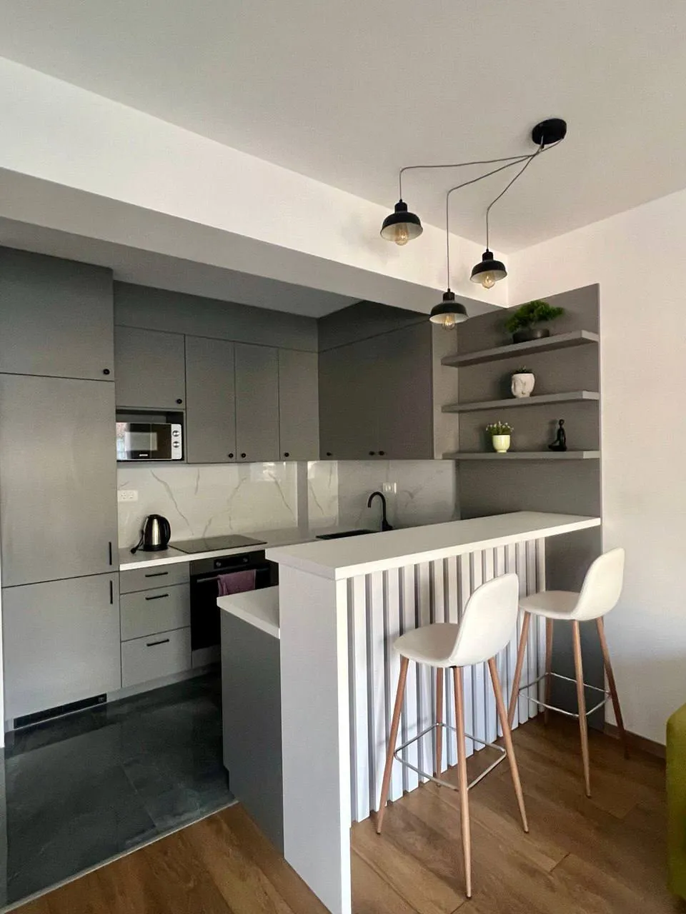 Modern kitchen with grey cabinets, a white bar counter, two white stools on wooden flooring and pendant lights.