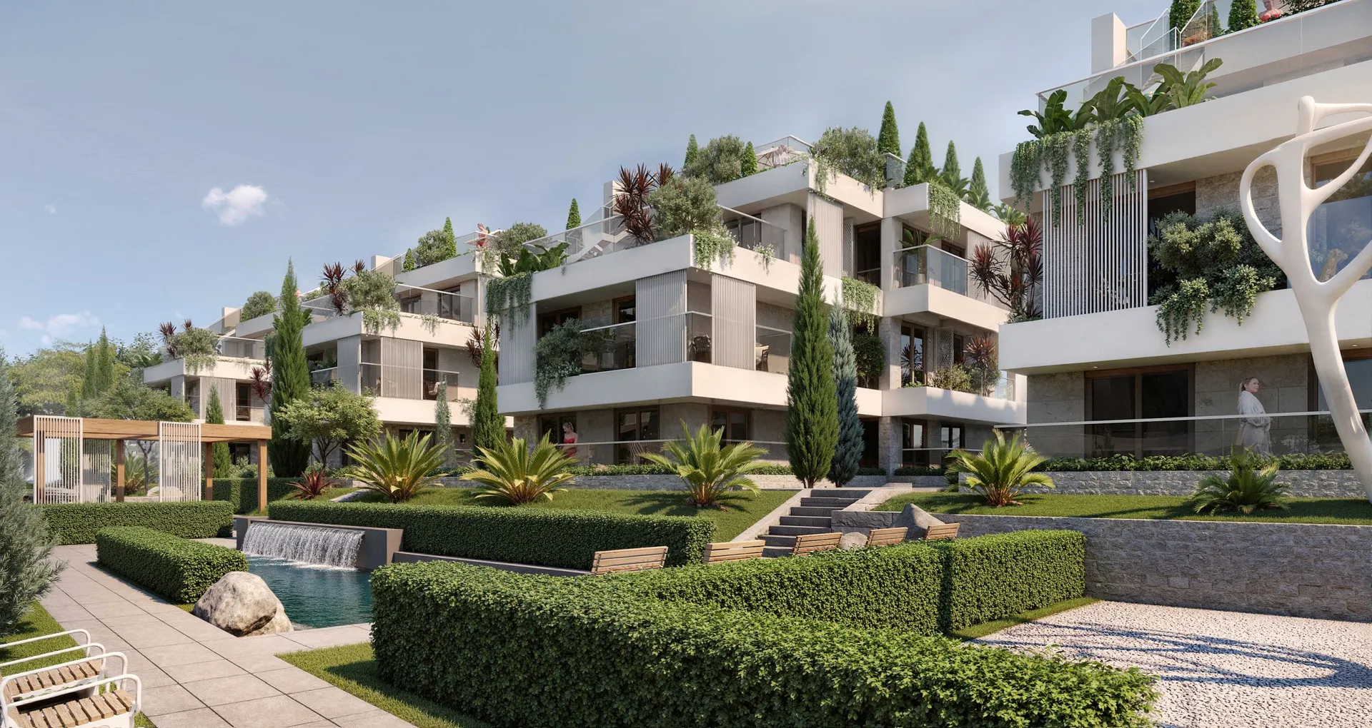 Modern residential complex featuring terraces, lush green gardens, a waterfall feature, and manicured hedges under a clear sky.