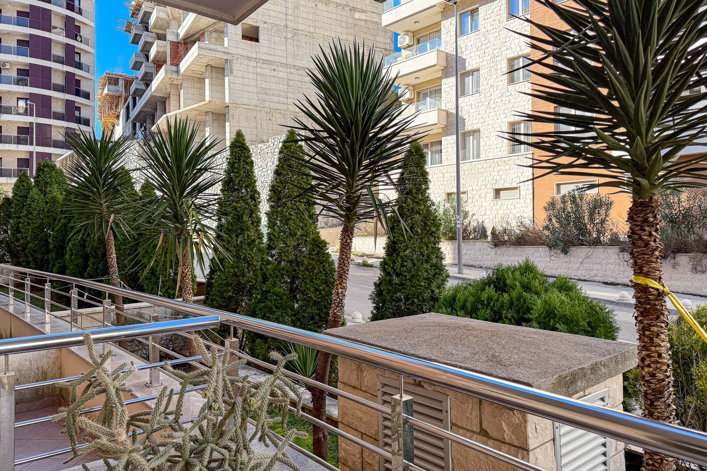View from the balcony of an apartment in Bečići, Montenegro: palm trees, thujas, modern residential buildings, and landscaped grounds.