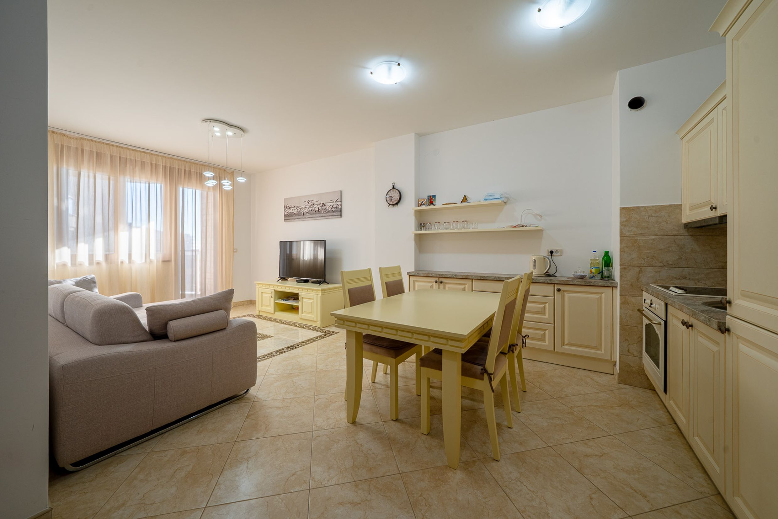 Living room with kitchen in an apartment in Bečići, Montenegro: light-colored furniture, dining area, tiled floor, and balcony access.