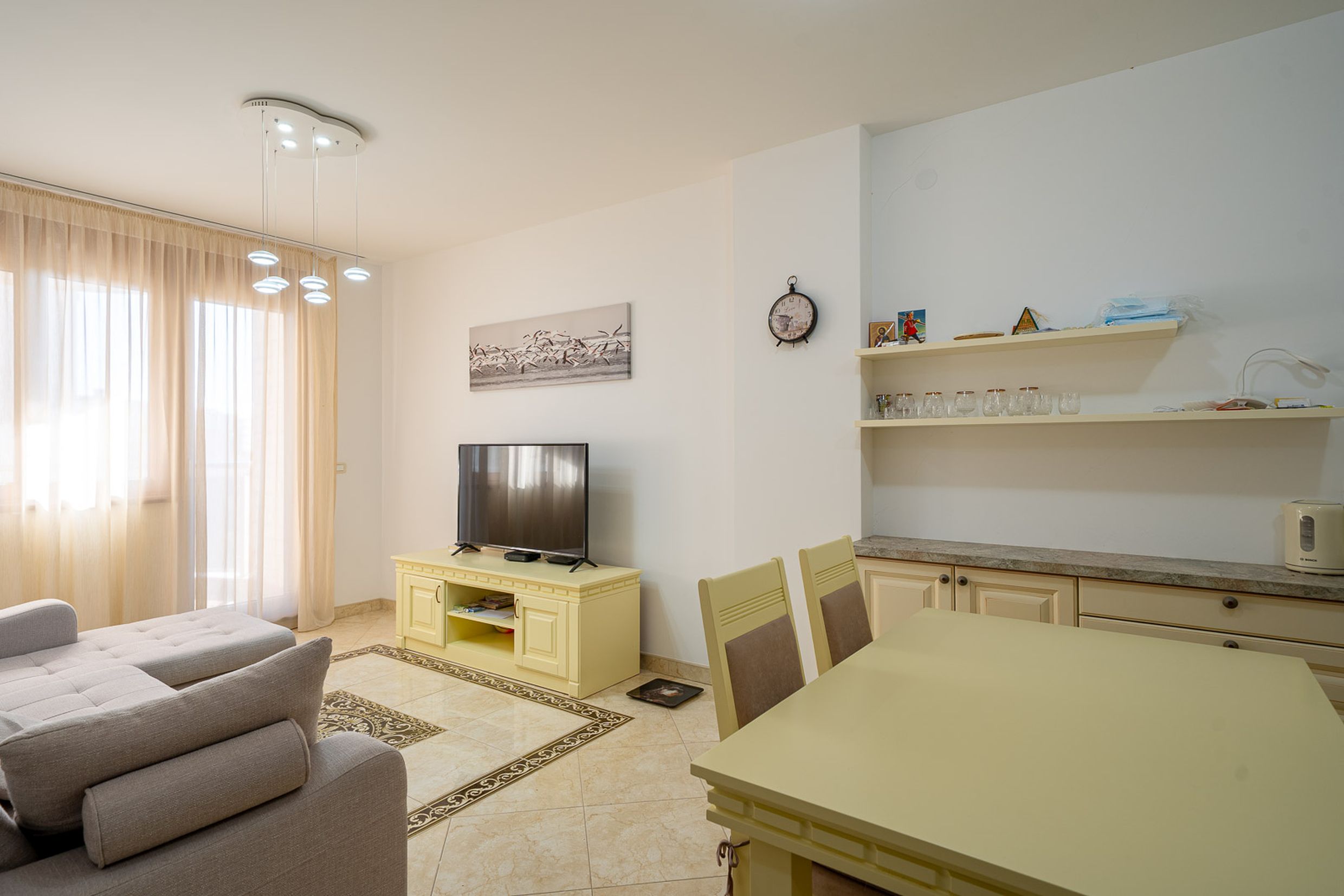 Bright living room with a dining area in an apartment in Bečići, Montenegro: plush sofa, TV stand, and tiled flooring.