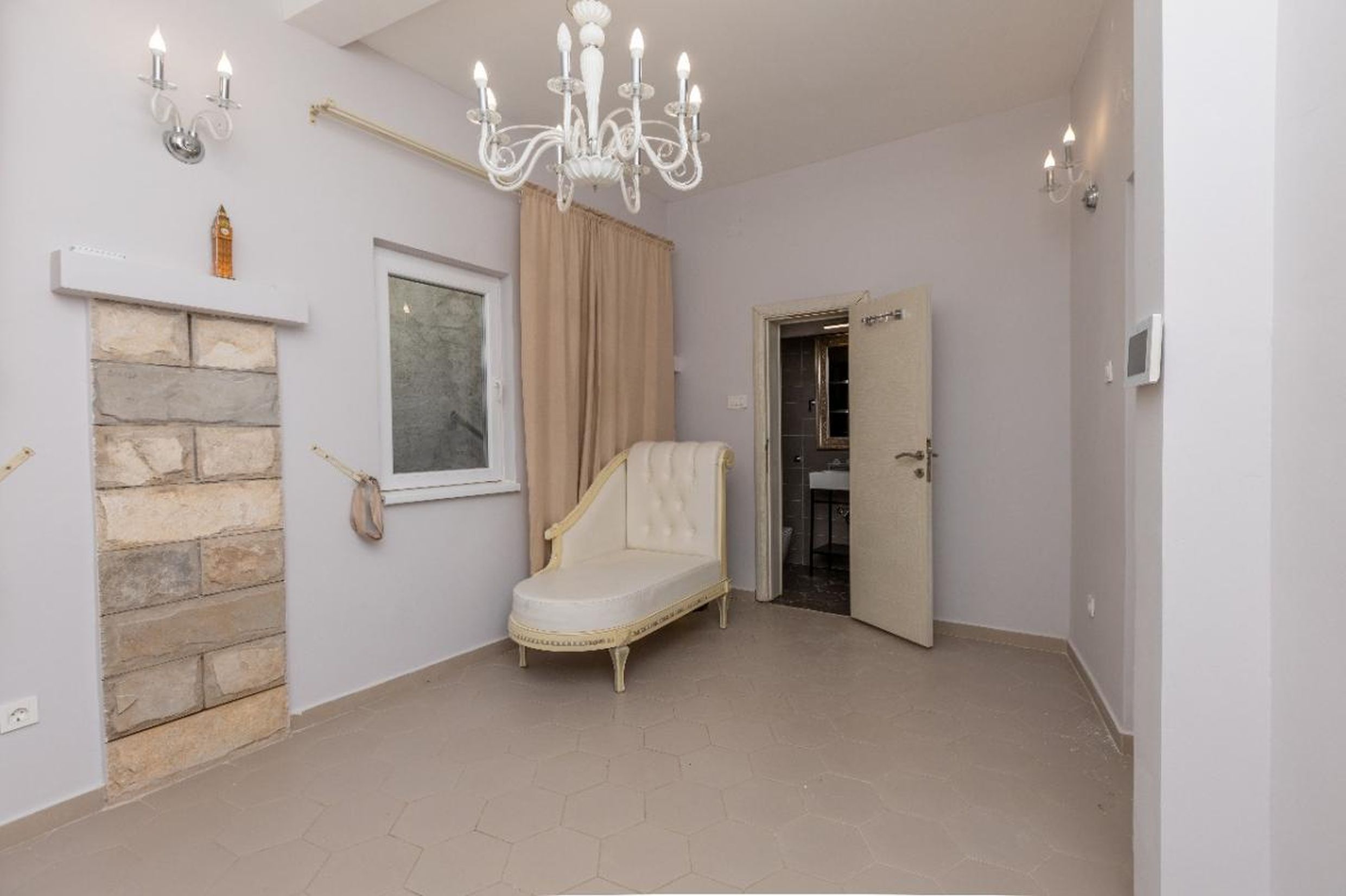 Living room in a house in Tivat, Montenegro: a classic chaise lounge, stone wall finish, chandelier, and entrance to the bathroom.