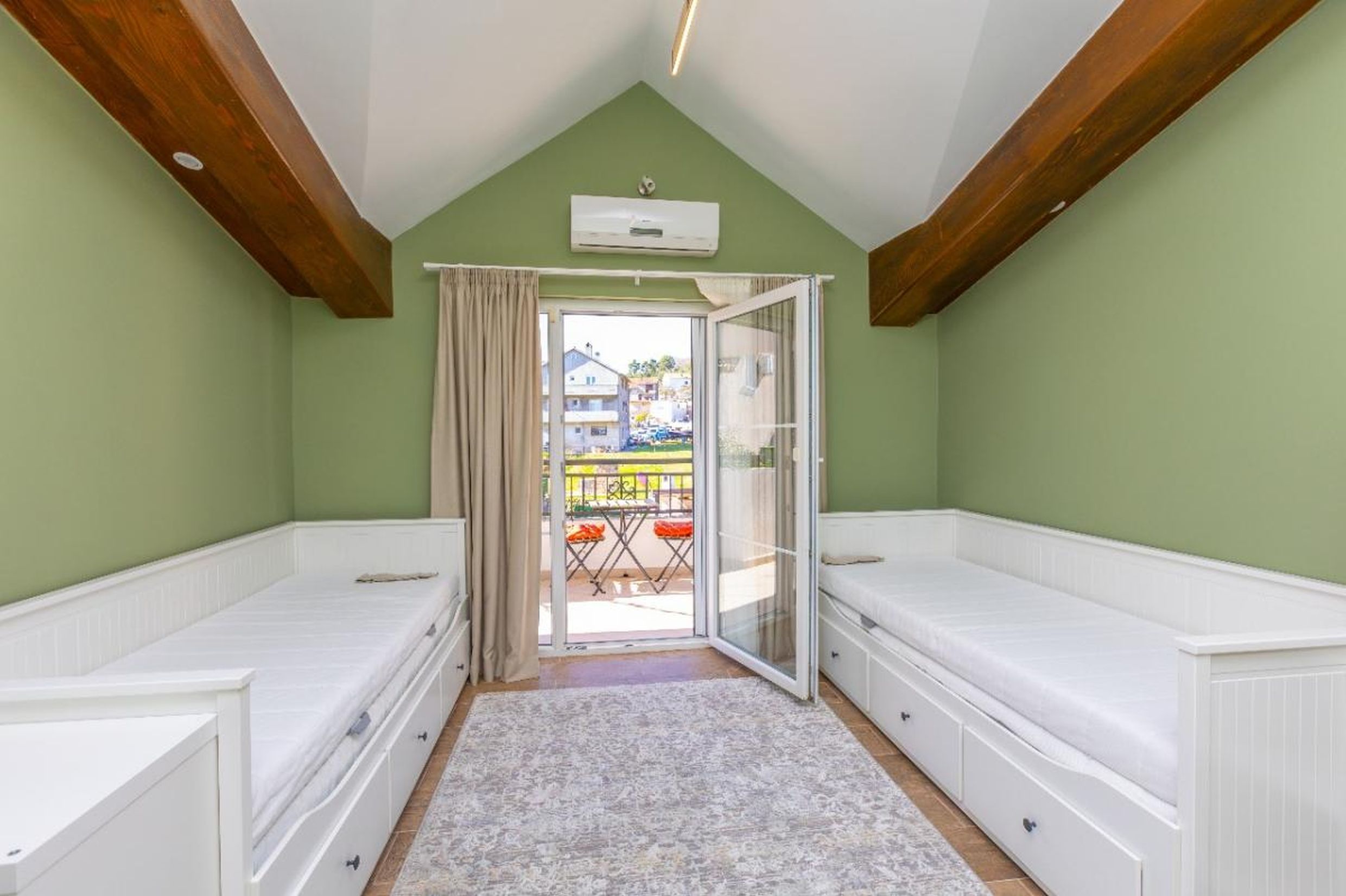 Attic bedroom in a house in Tivat, Montenegro: two white beds, wooden beams, access to a balcony with a view.