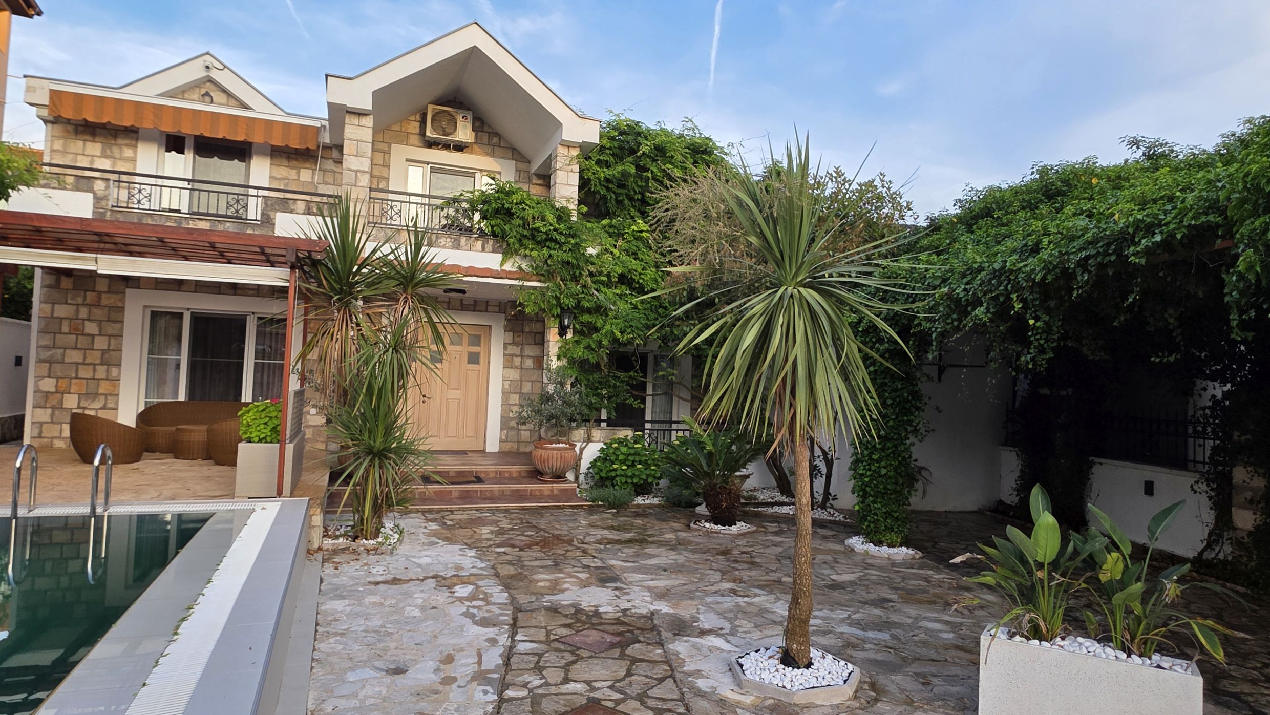 Two-story house in Tivat, Montenegro: stone facade, terrace with a pool, palm trees, and a well-kept garden.