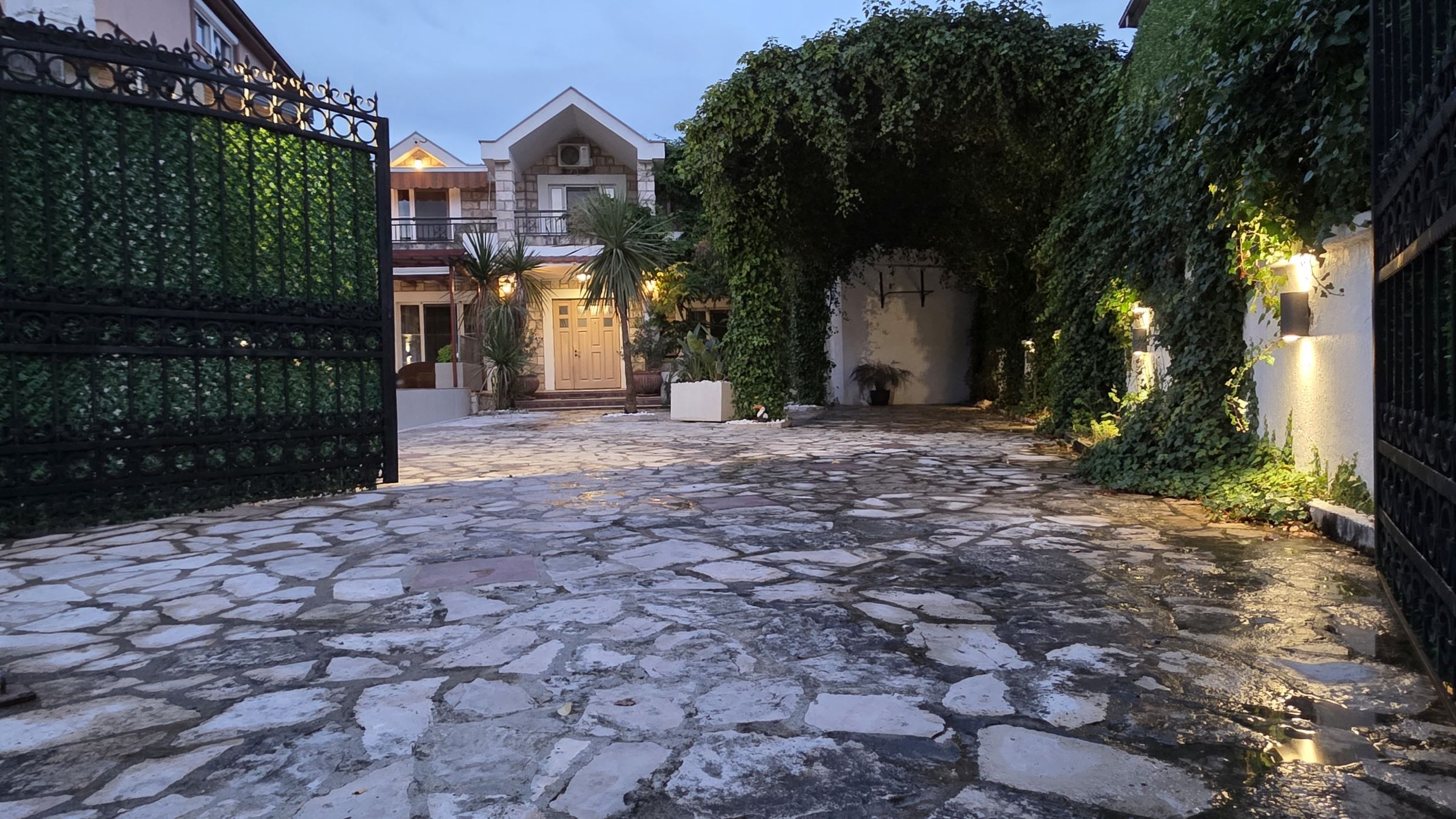 Courtyard of a private house in Tivat, Montenegro: a stone-paved area, wrought-iron gates, and a green arch of climbing plants.