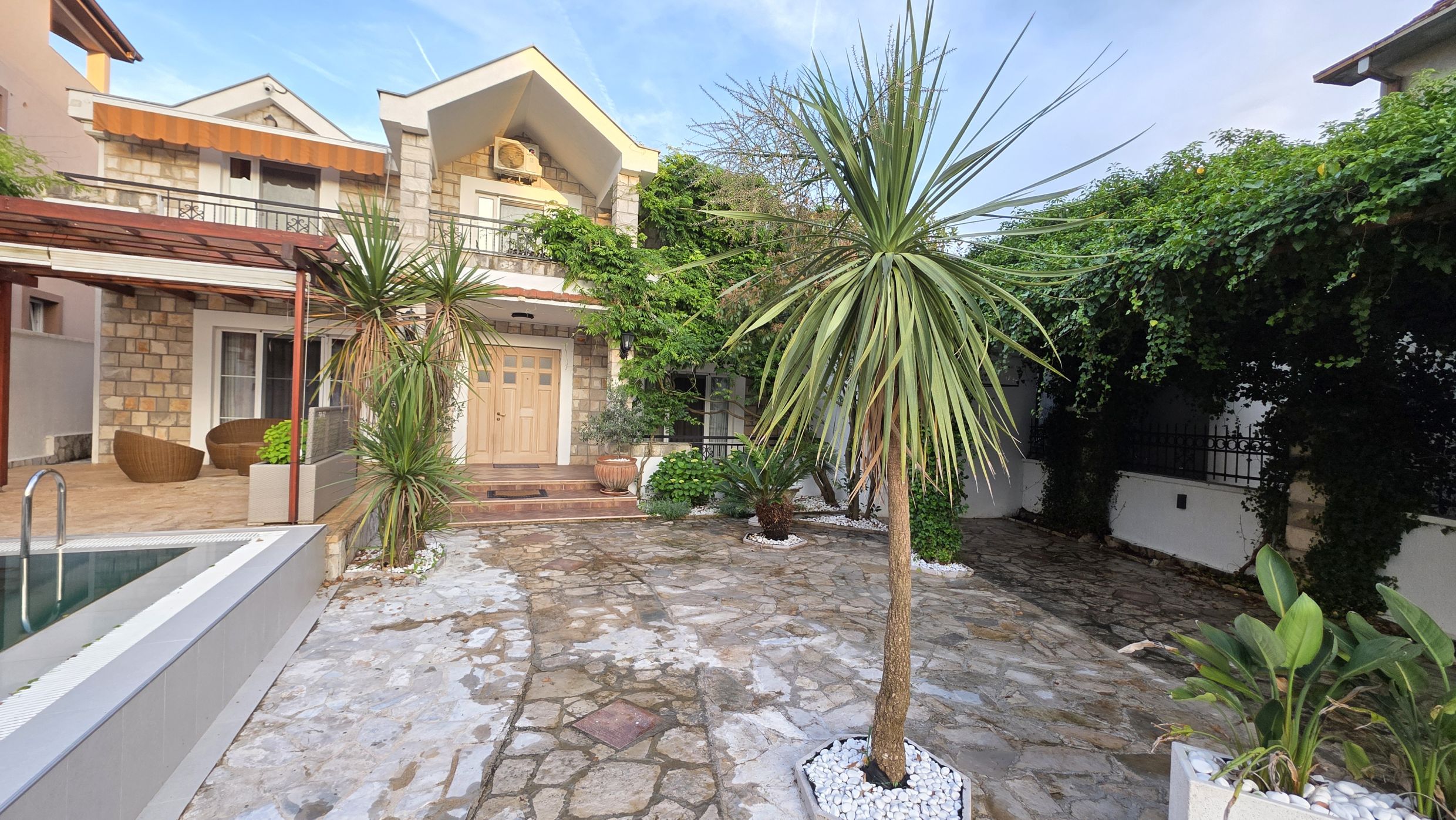 Two-story house in Tivat, Montenegro: stone facade, terrace with rattan furniture, pool, and a well-kept garden.