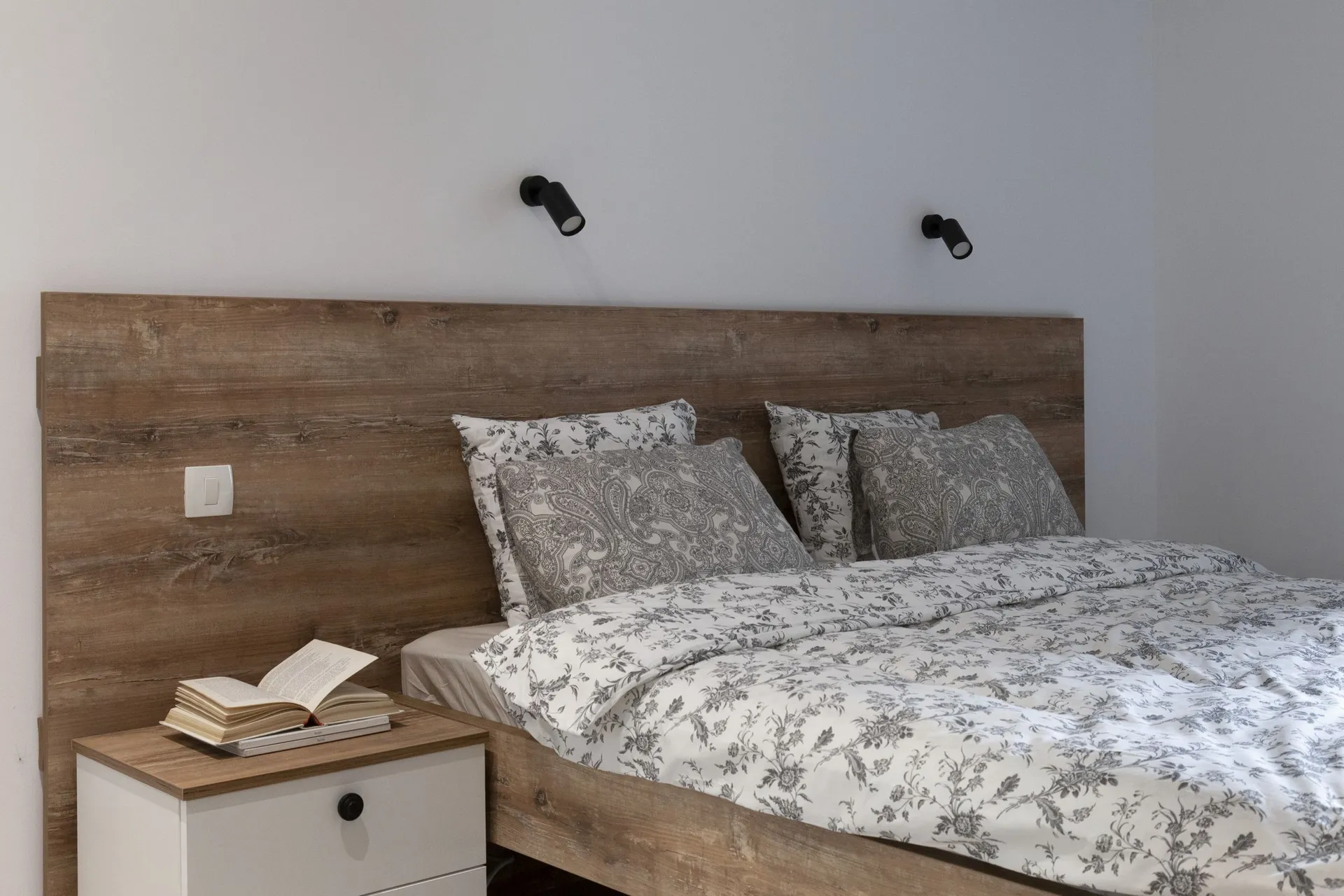 Modern bedroom featuring a rustic wooden headboard, grey floral bedding, and black wall-mounted spotlights above the bed.