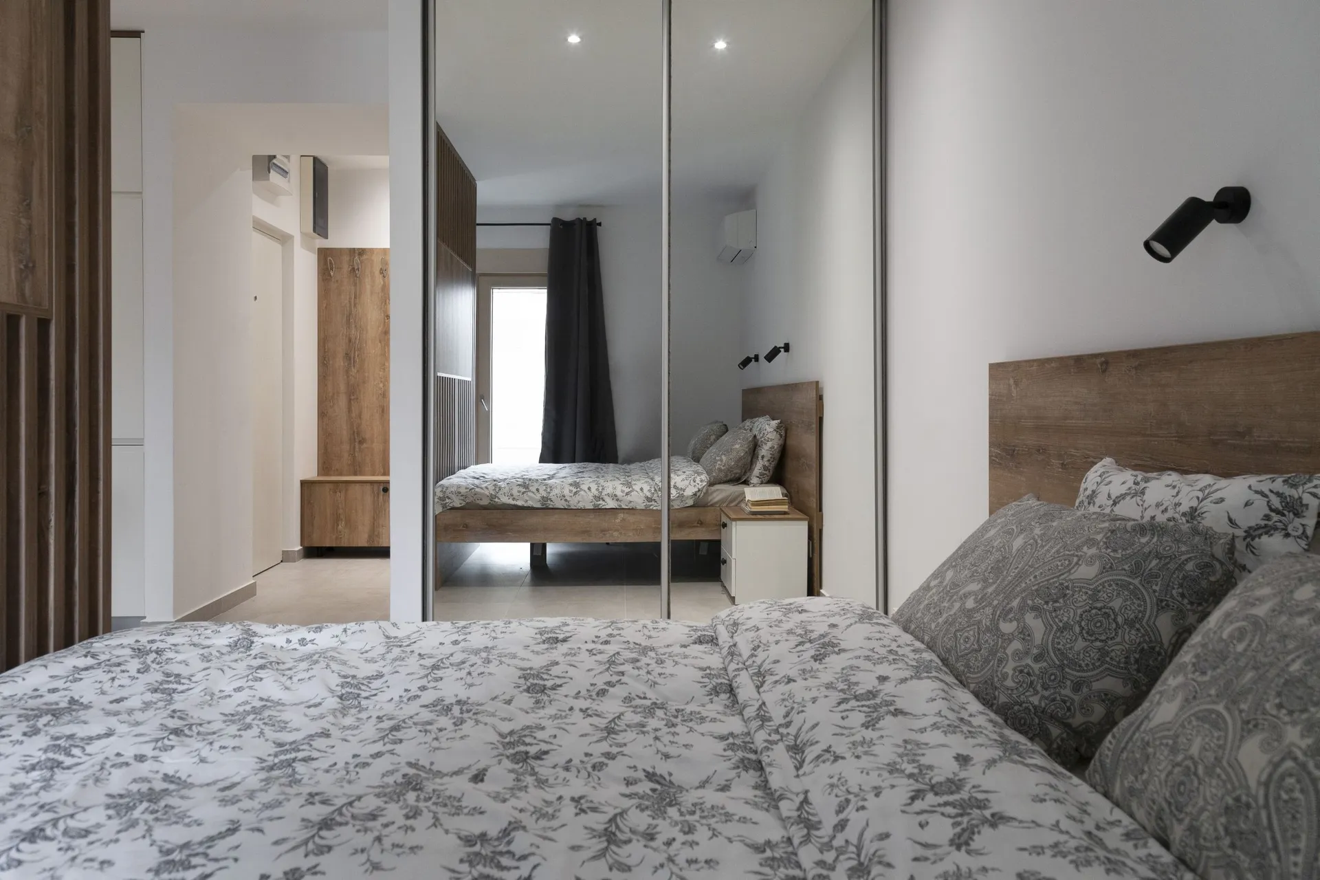Modern bedroom featuring a wooden bed with floral bedding and a large mirrored wardrobe reflecting the room.