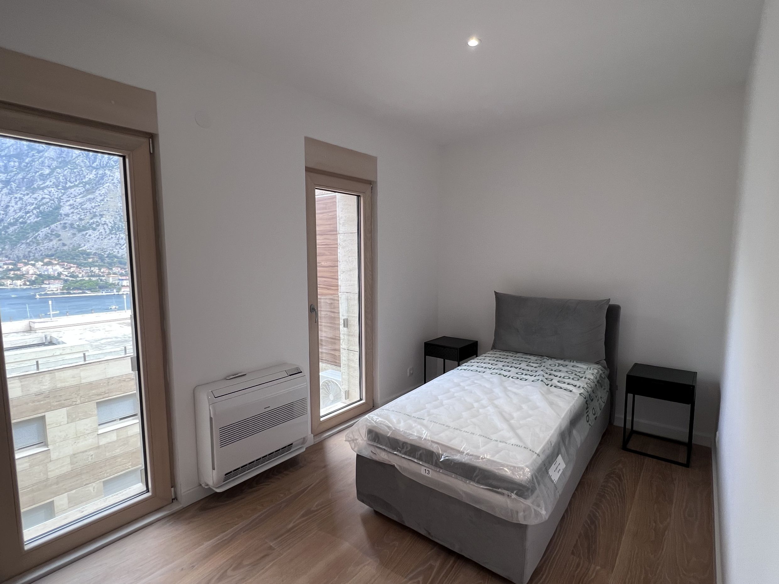 Bedroom in an apartment in Kotor, Montenegro: bed with mattress, black nightstands, panoramic view of Kotor Bay and the mountains.