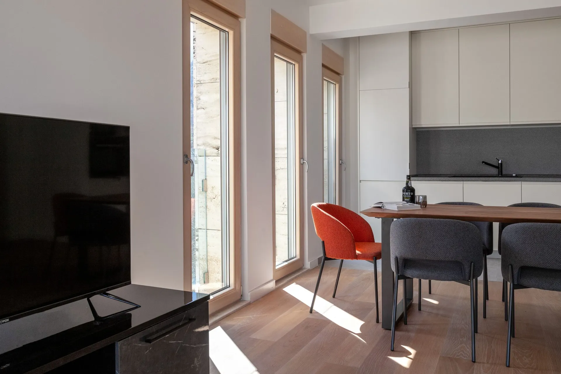 Modern living room featuring a TV, wooden dining table, bright orange chair, and an open kitchen area visible in the background.