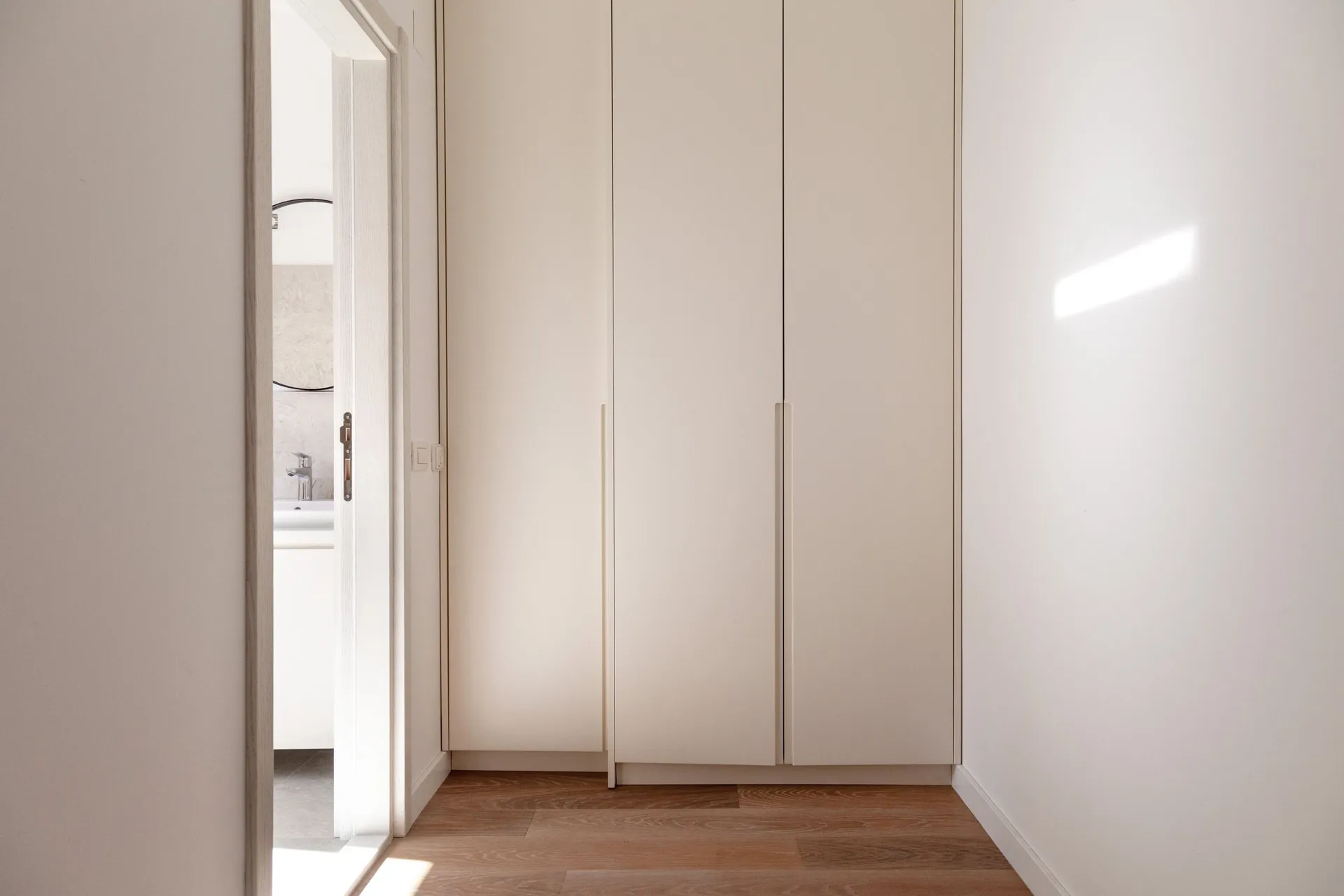 Bright hallway with white built-in wardrobes and wooden flooring. An open door leads to a bathroom visible in the background.